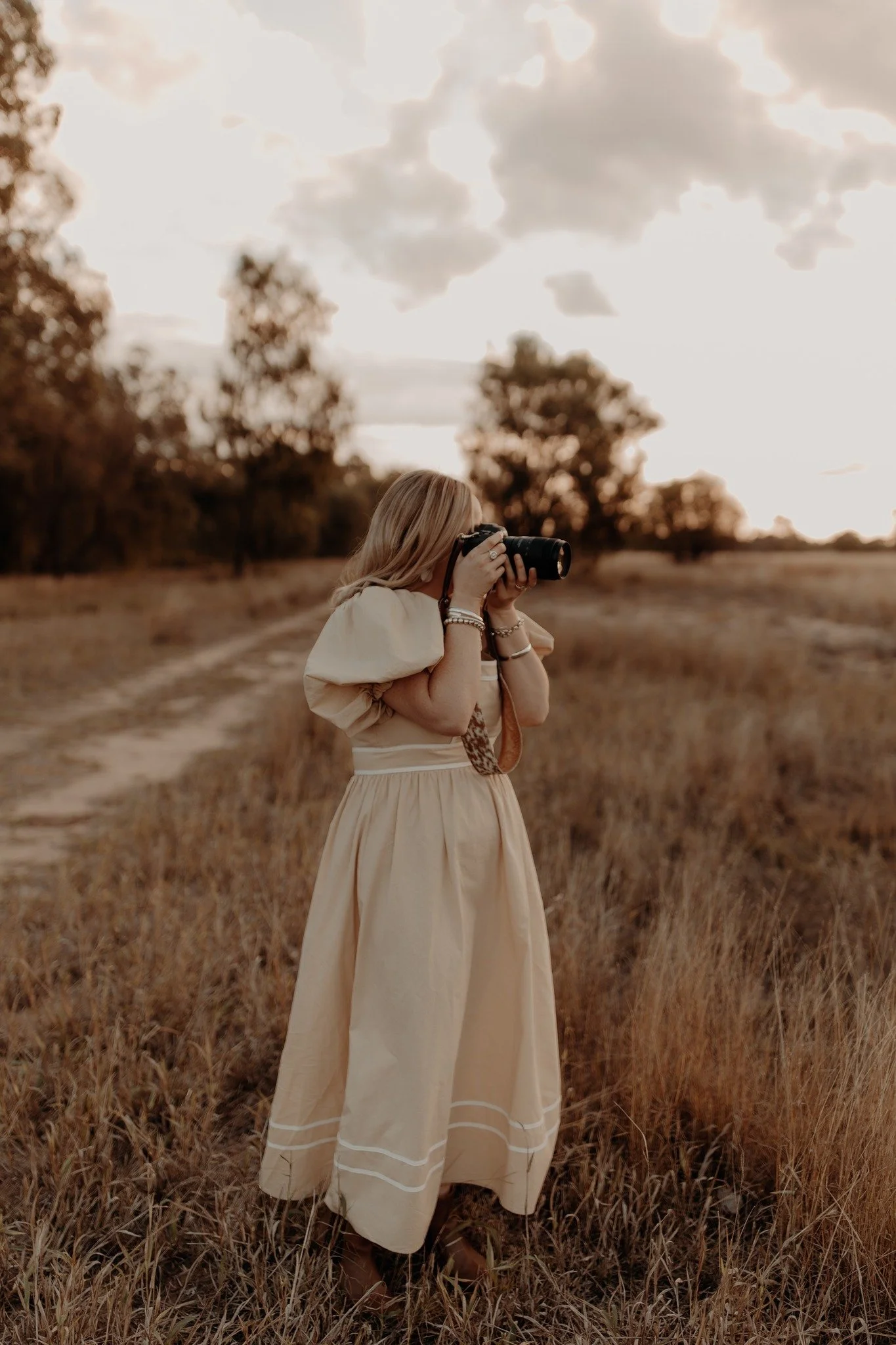 A woman with blonde hair, wearing a beige dress, standing in a field of dry grass, holding a camera up to her face, capturing a photo during sunset with a cloudy sky.