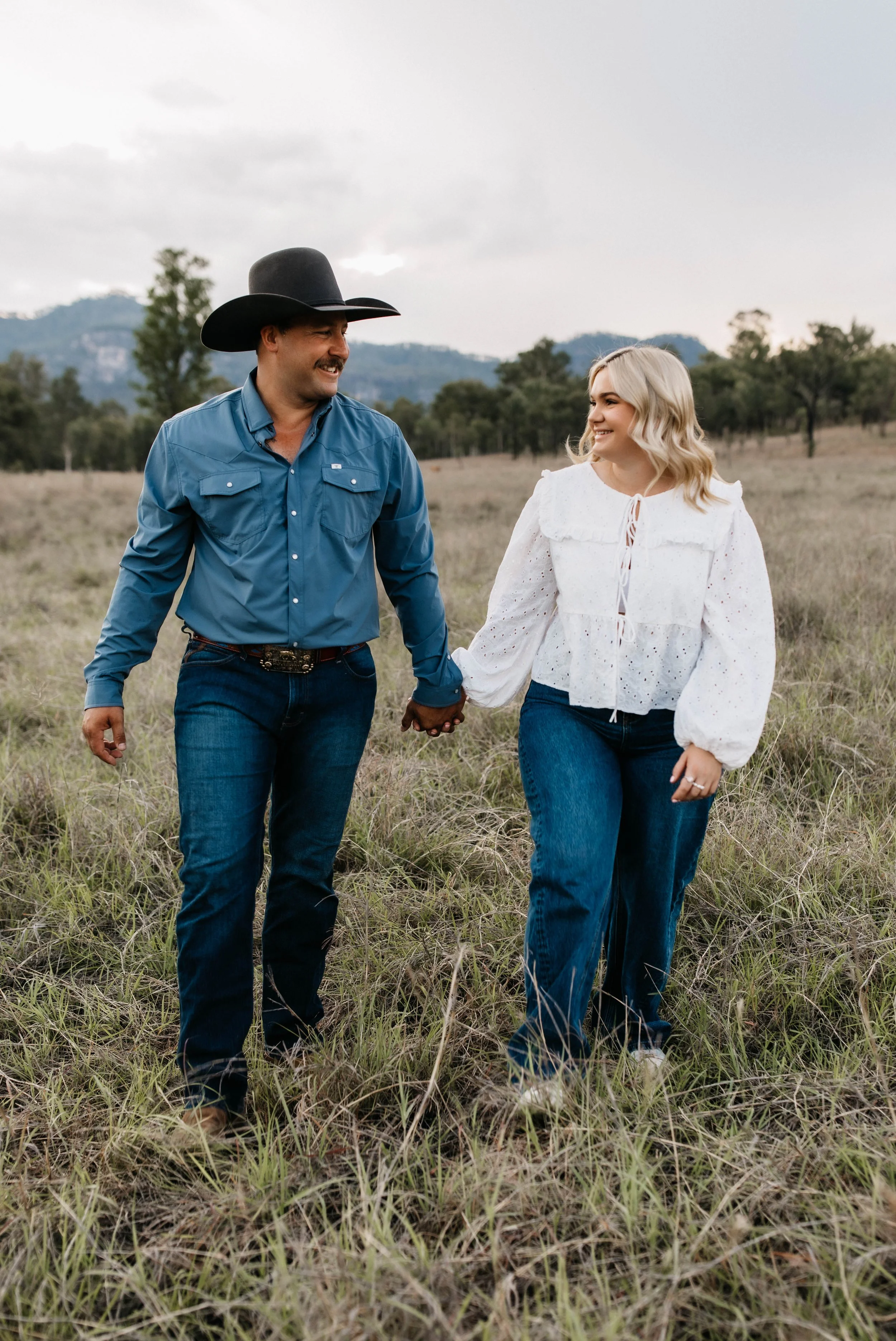 A man and woman walking hand in hand through a grassy field, smiling at each other, with trees and mountains in the background.