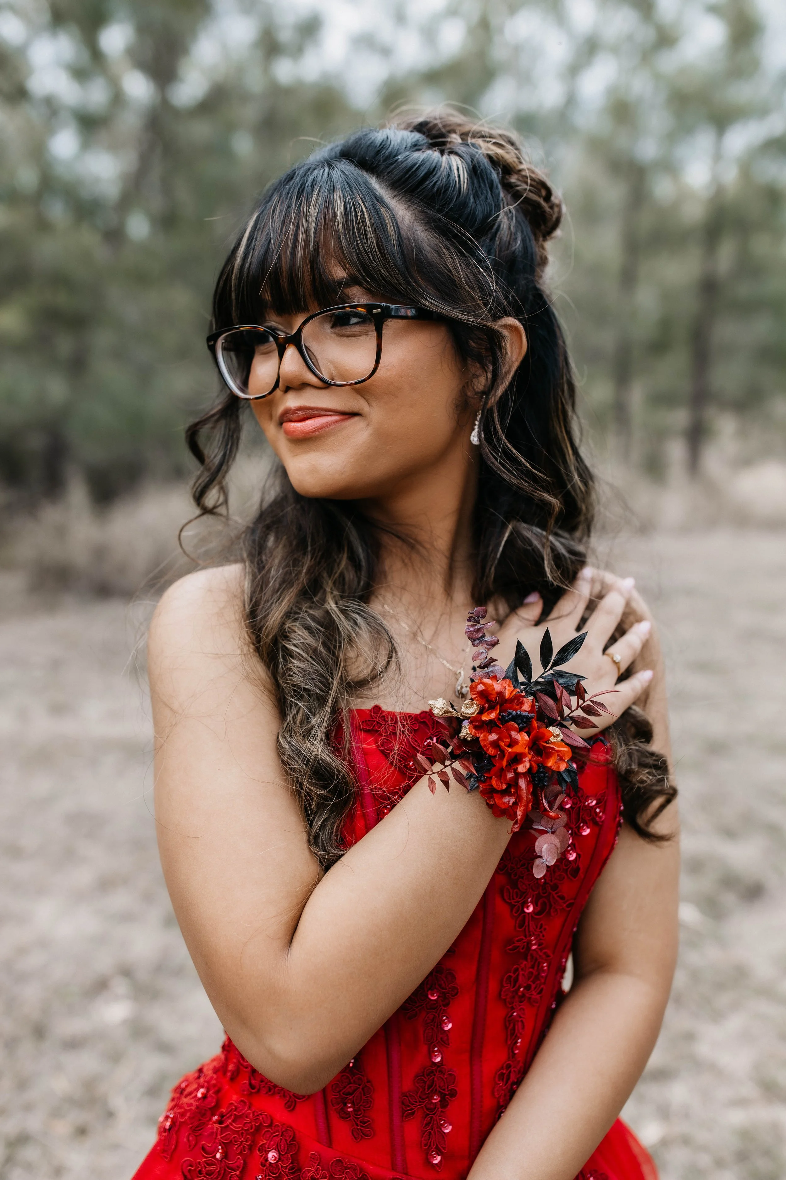 A woman with glasses and dark hair styled in waves and an updo, wearing a red dress with floral embroidery, holding a bouquet of red and dark purple flowers, amid a blurred outdoor background of trees.