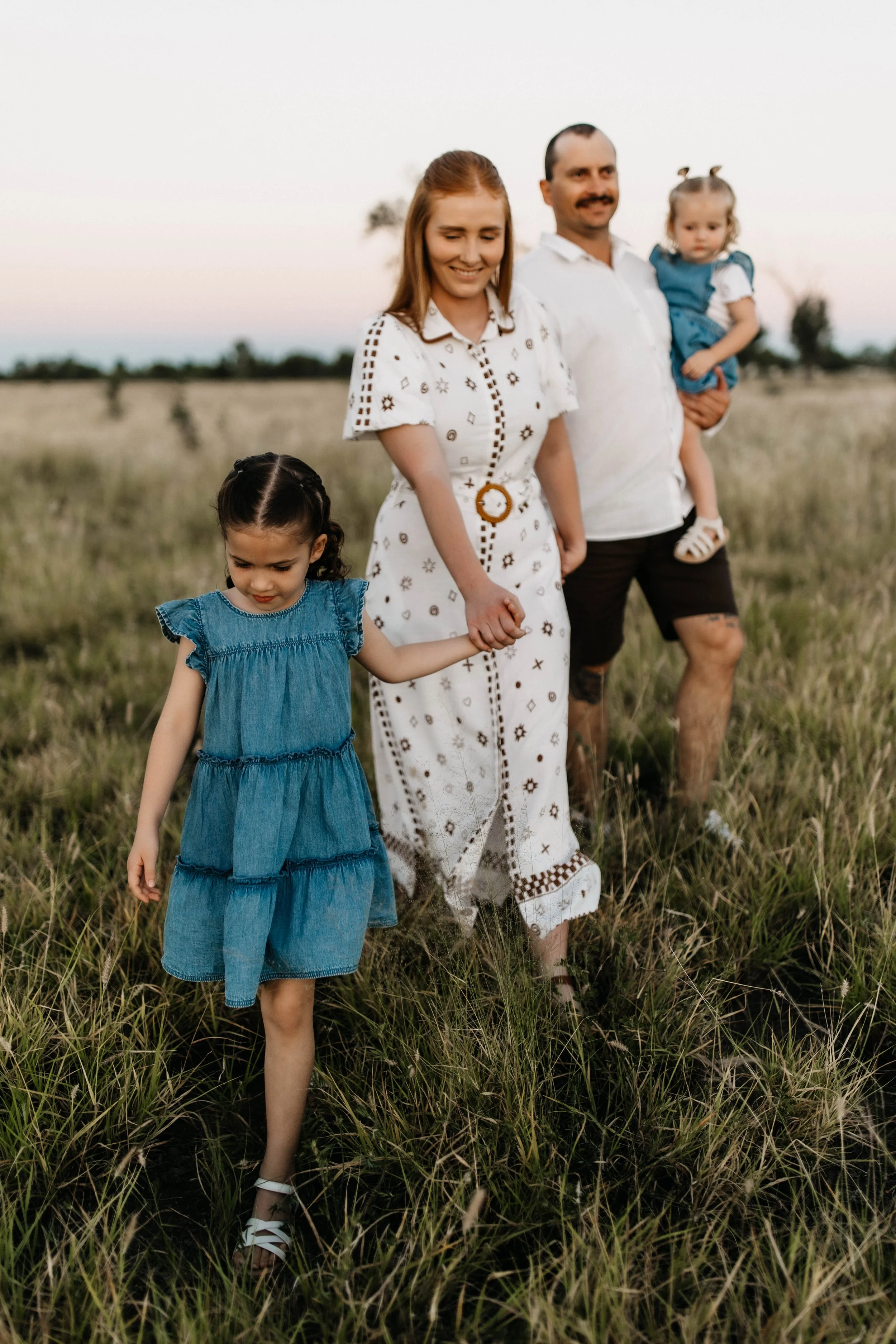 A family of four walking through a grassy field during dusk. The mother holds hands with a young girl, and the father carries a toddler girl. The sky is light pink and blue, with trees in the background.