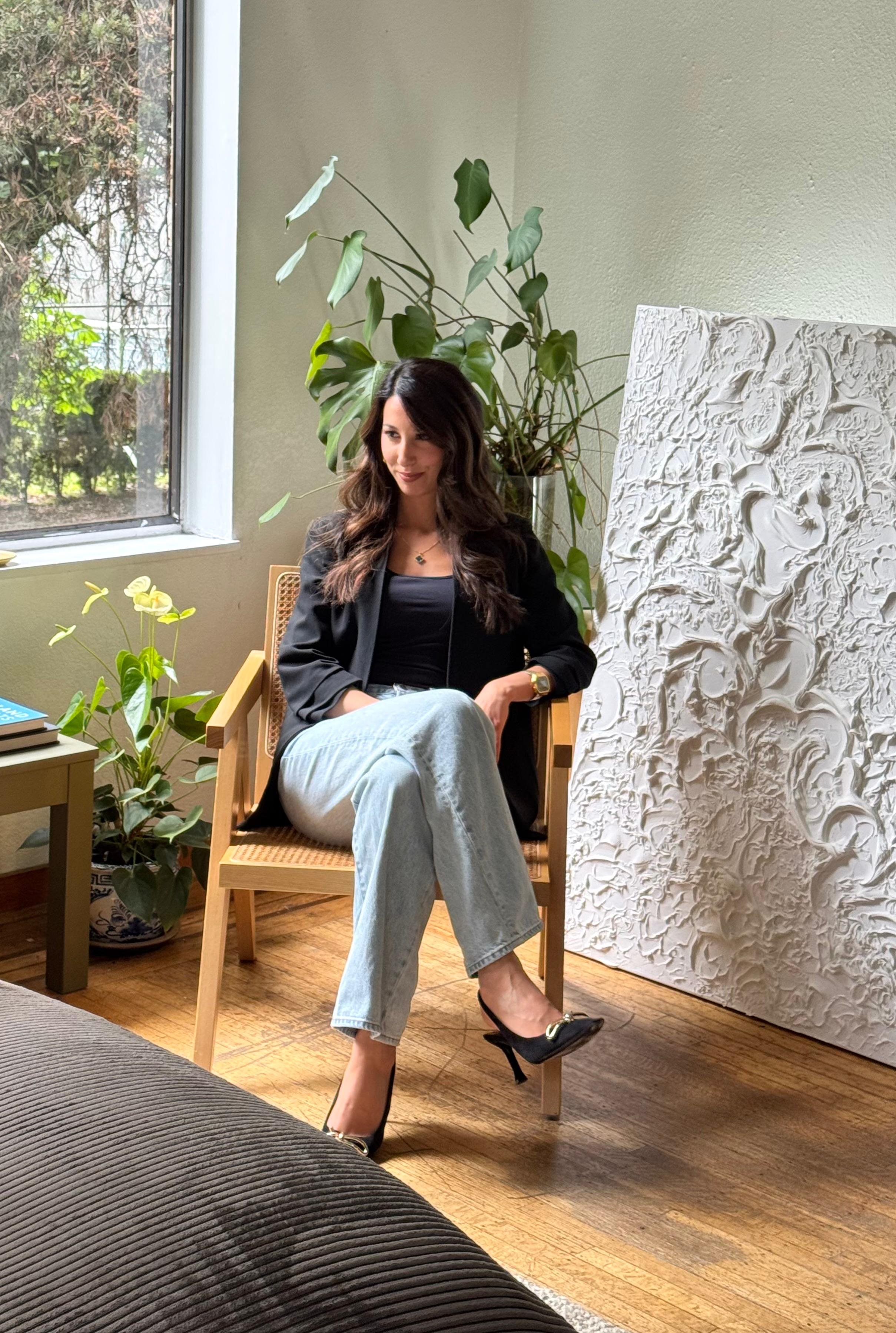 A woman with long dark hair sitting on a wooden chair indoors, wearing a black blazer, black top, light blue jeans, and black high heels, with a large green houseplant and abstract white textured artwork behind her, near a window letting in natural light.