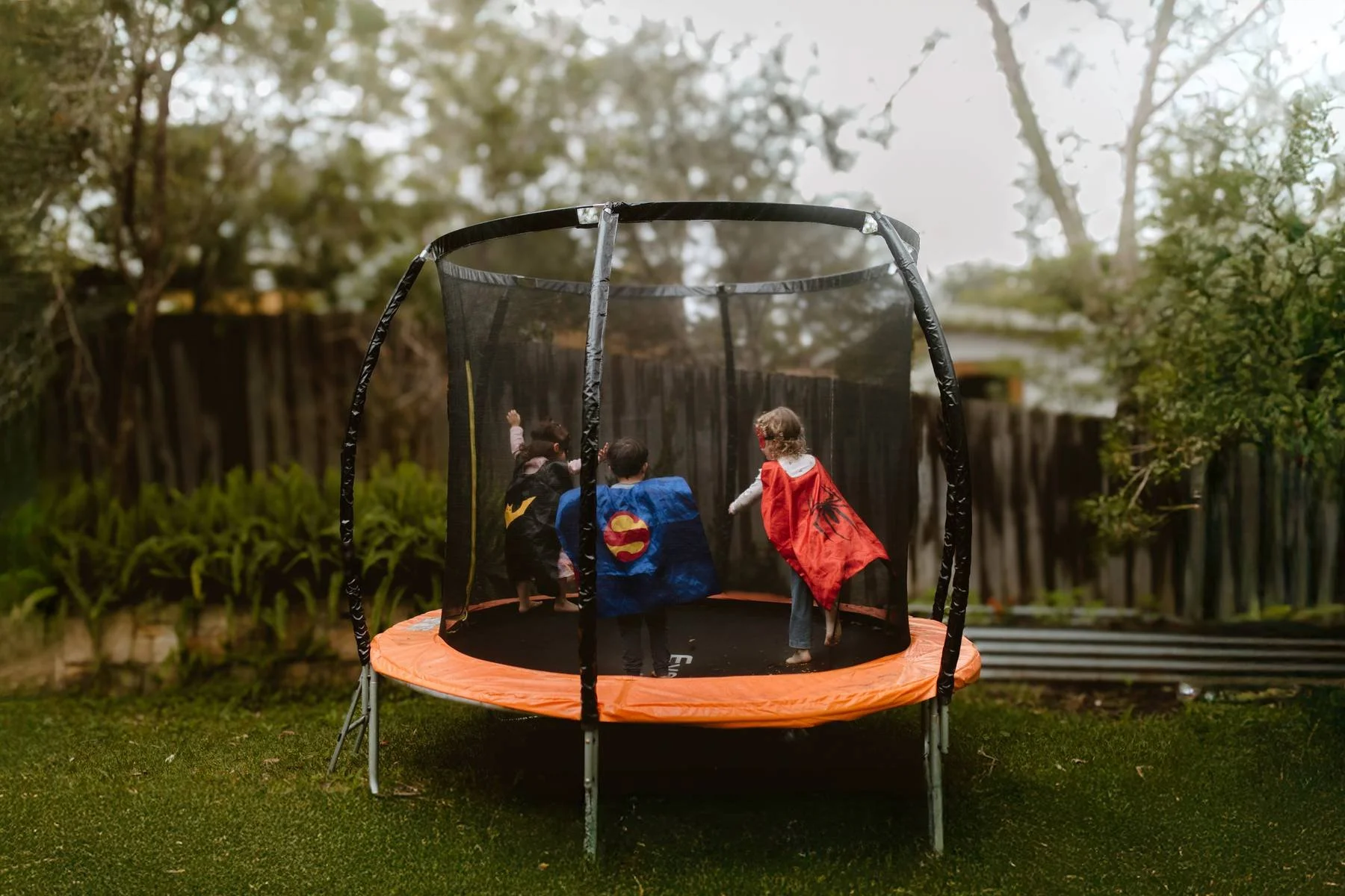 Three children dressed in colourful superhero capes and masks, joyfully jumping mid-air on a backyard trampoline.