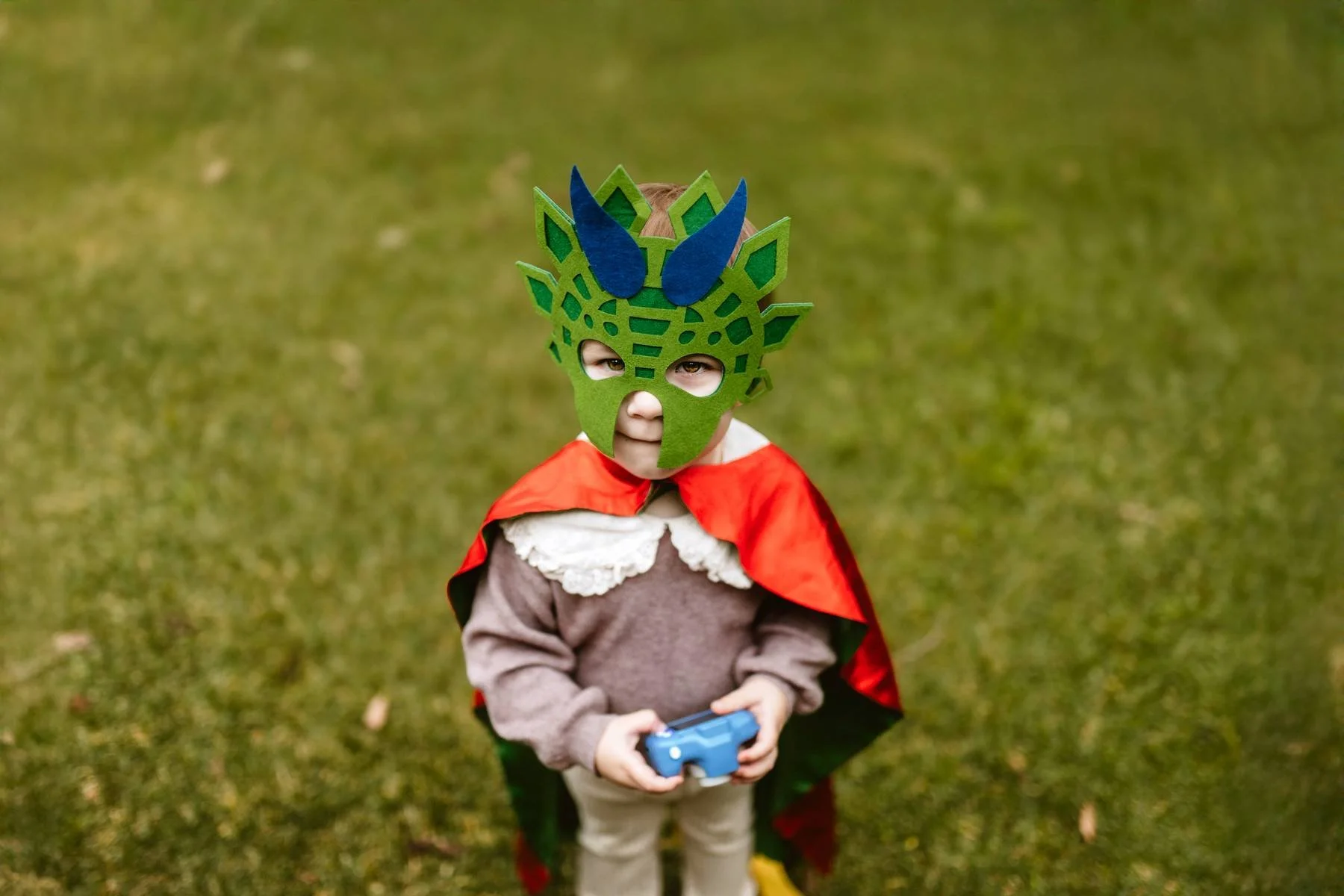 Young child standing holding a toy camera wearing a green dragon mask and red cape