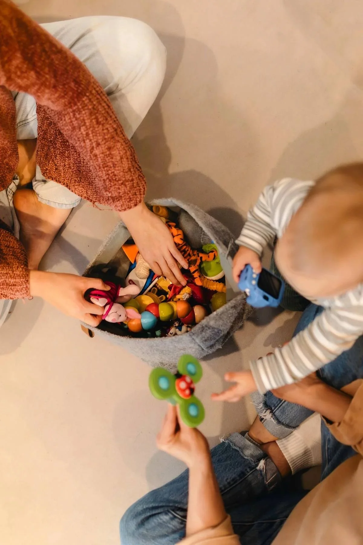 Two children and an adult are playing with a large gray basket filled with colorful toys, including stuffed animals, plastic balls, and toy figure. The children are reaching into the basket, one holding a toy and the other reaching for more toys.
