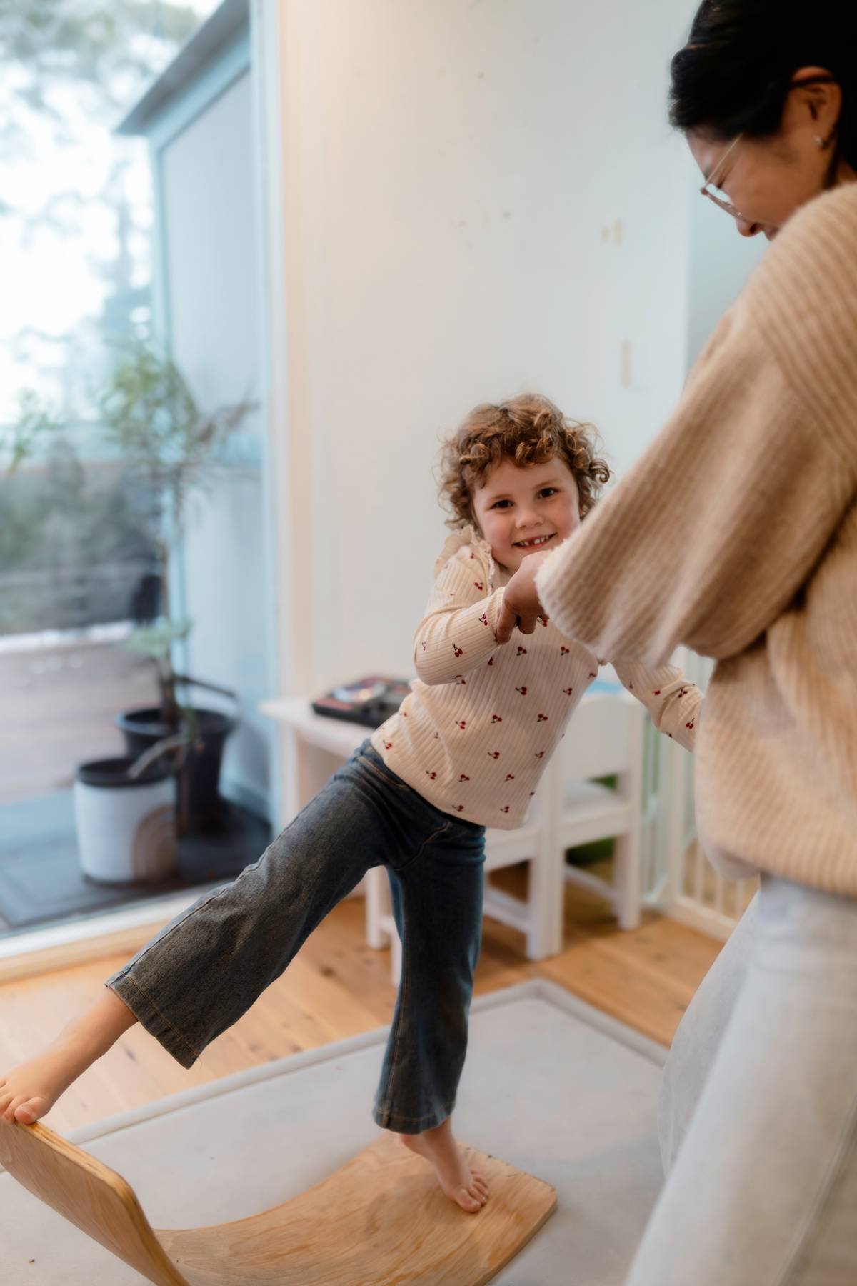 Smiling girl on a wooden balance board holding hands with a woman facing away and smiling at the girl.