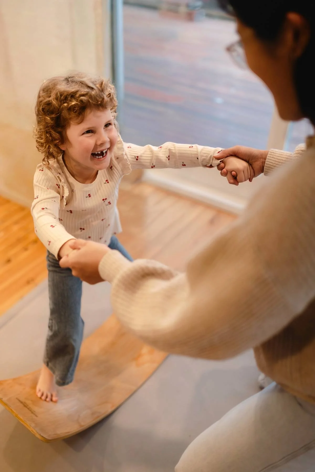 Child on a wooden wobble board and smiling whilst holding Occupational Therapist's hands.