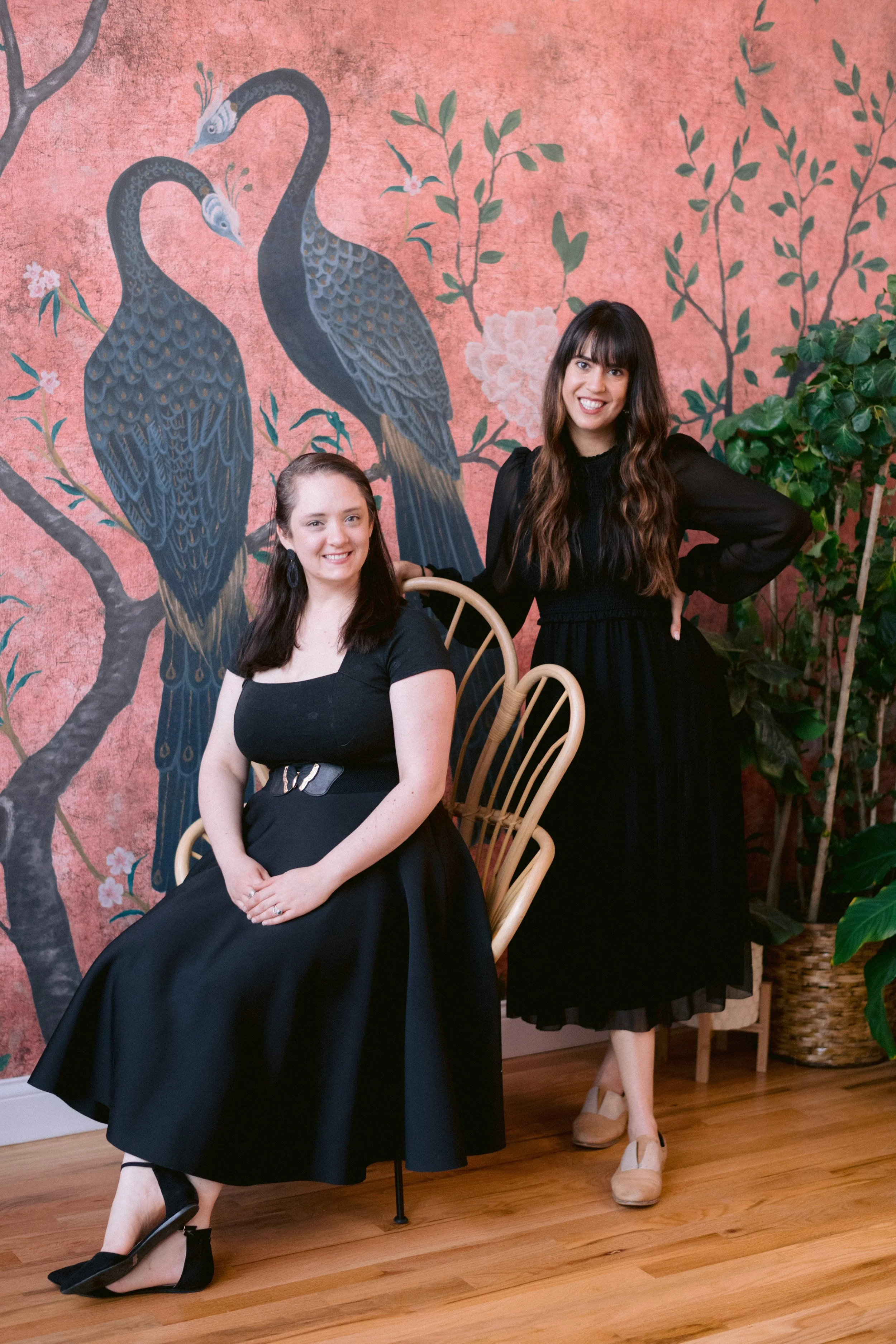 Two women posing in front of a pink wall with a painted mural of two cranes and branches with flowers. One woman is sitting on a chair and the other is standing beside her.