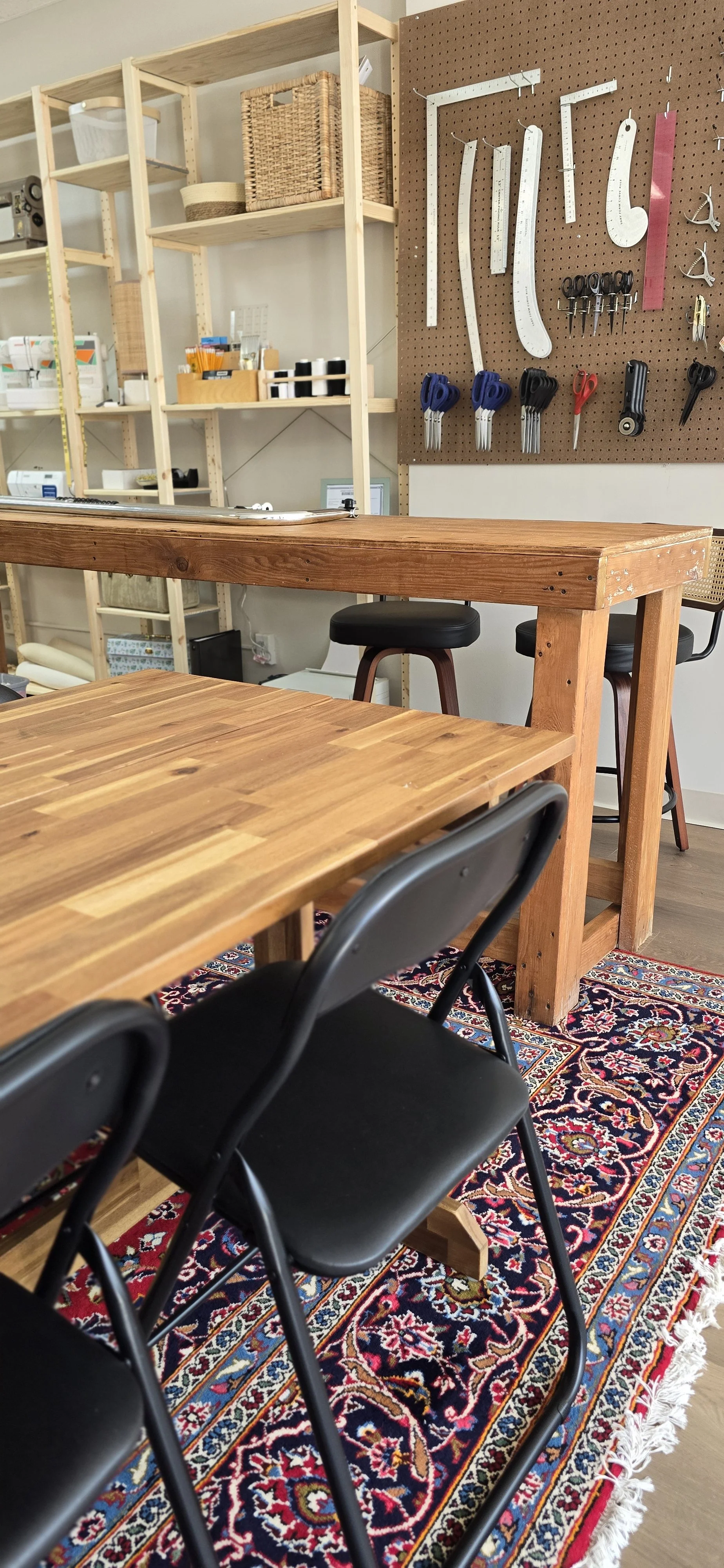 Wooden tables and black folding chairs, sewing tools on pegboards and a shelf full of sewing machines