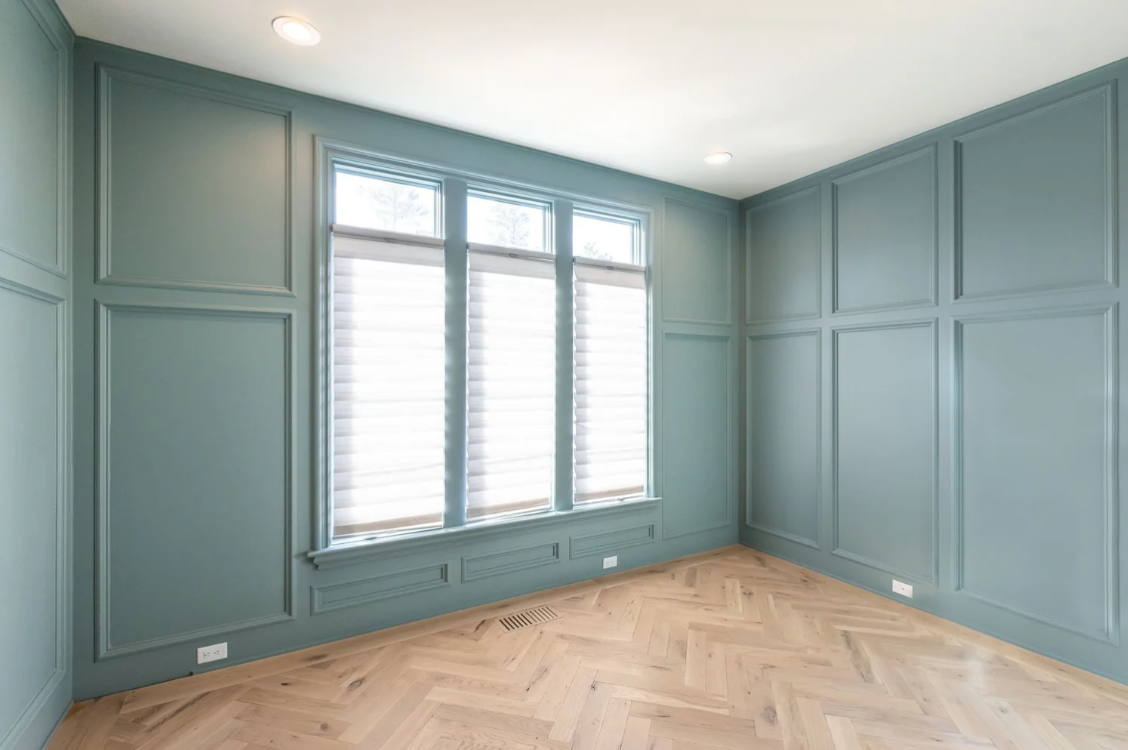 Empty room with teal walls, hardwood herringbone floor, large windows with white blinds, and recessed lighting.