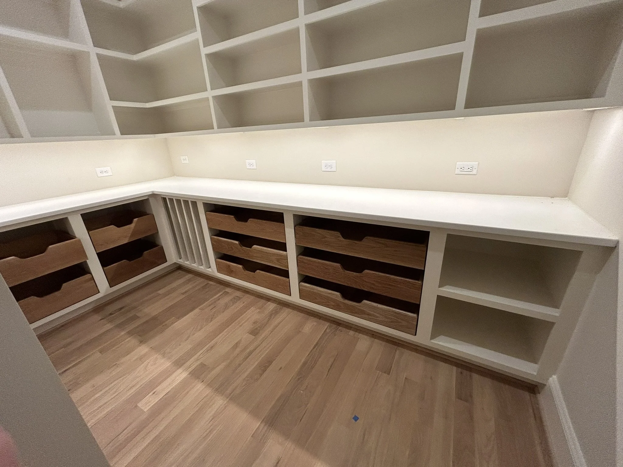 Empty built-in white shelving unit with open compartments and drawers with wooden fronts, set in a room with hardwood flooring, multiple electrical outlets on the wall, and no other furniture or decorations.