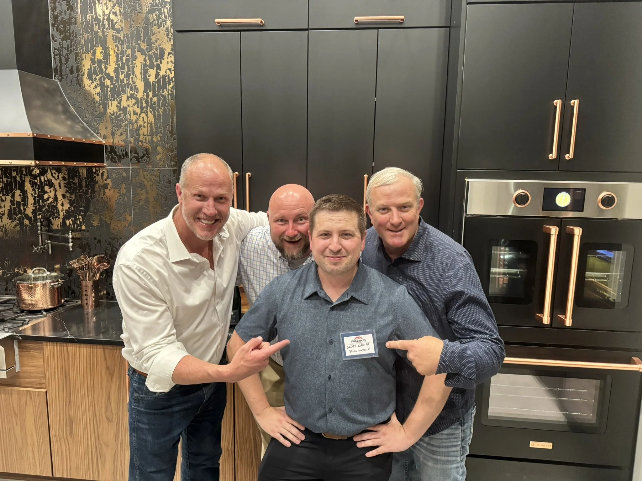 Four men smiling and posing together in a modern kitchen. One man is holding a name tag that reads 'Scott Camp,' with a subtitle in smaller text beneath. The kitchen features black and copper cabinets, a stainless steel oven, and copper handles and fixtures.
