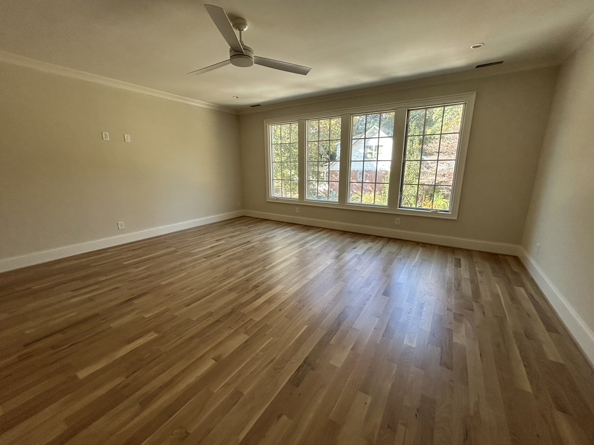 Empty room with large window, wooden floor, ceiling fan, and beige walls.