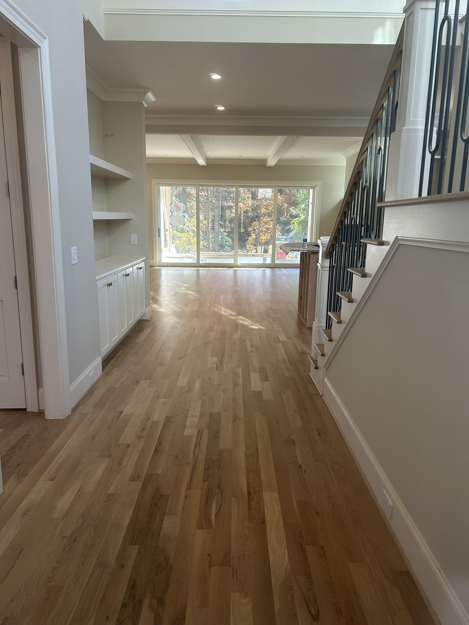 Living room with hardwood floors, large sliding glass door showing trees outside, built-in shelves, staircase with wooden steps and metal railing.