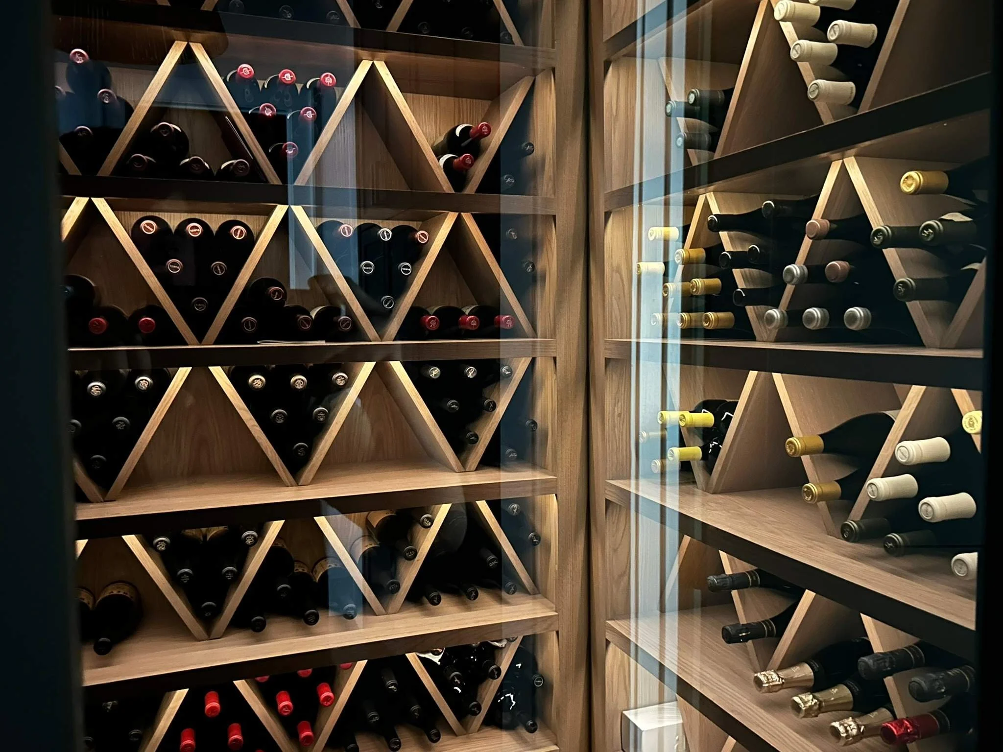 A wine cellar with wooden racks holding bottles of wine stored horizontally, with glass doors reflecting light.