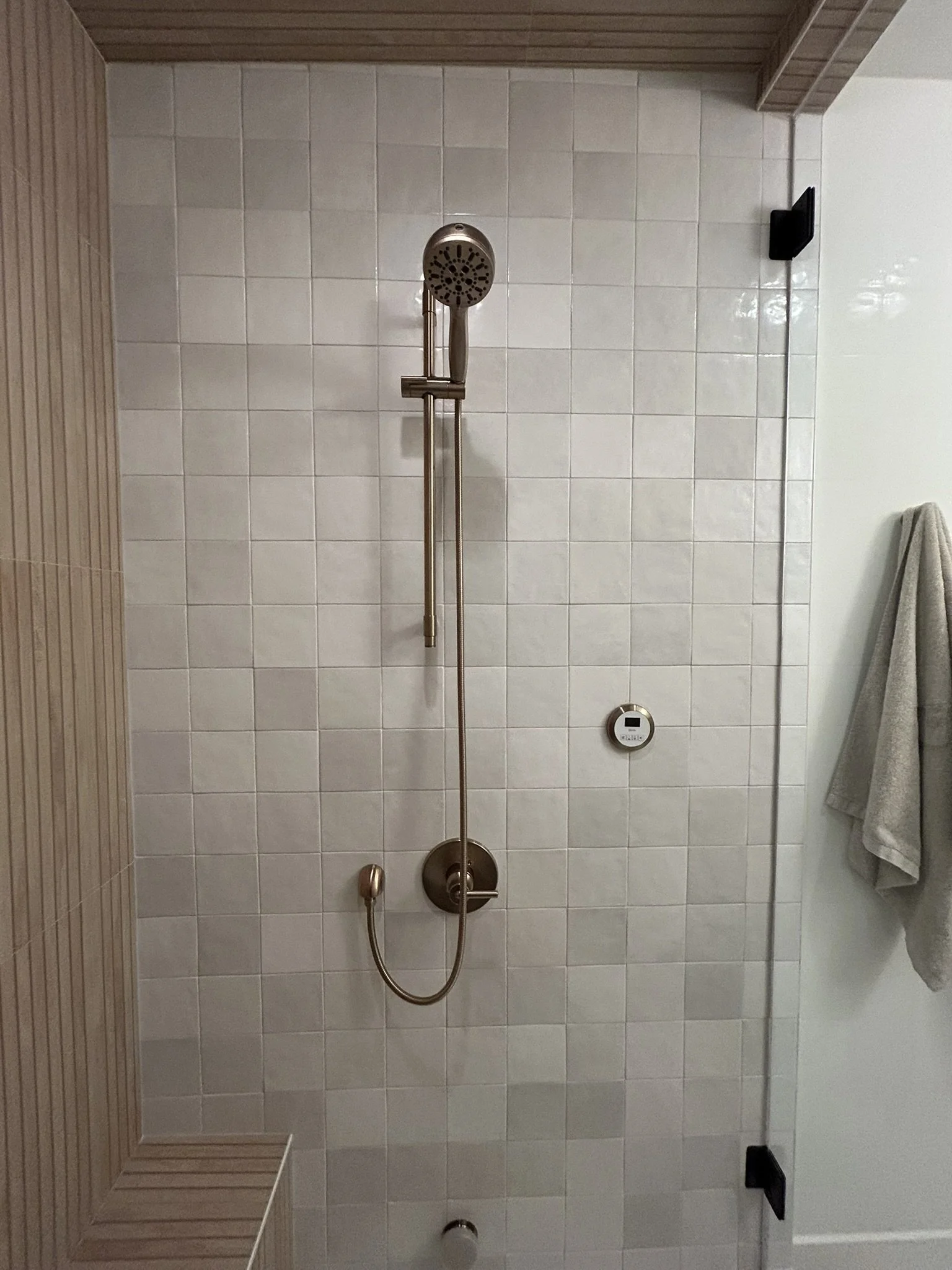 A shower area with white tiled wall, a brass showerhead, a matching brass handle, a black shelf on the wall, and a gray towel hanging on a black hook on the white wall.