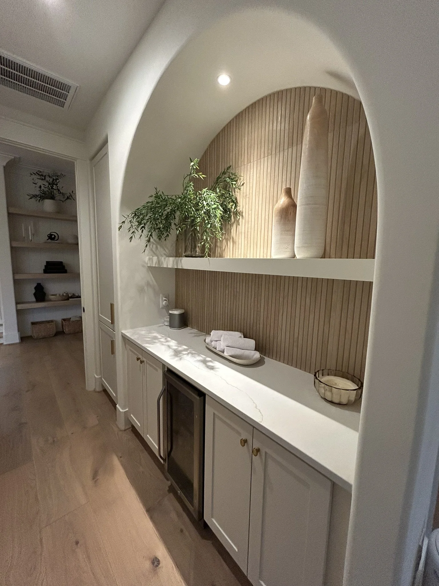 A white sideboard with closed cabinets, topped with a marble surface, positioned against a wall with wooden paneling. Decor includes two large beige vases, a small potted plant, a black speaker, and folded towels. To the left, there is a doorway leading to a room with open shelves holding various decorative items. The floor is made of light-colored wood, and the ceiling has a recessed light fixture.