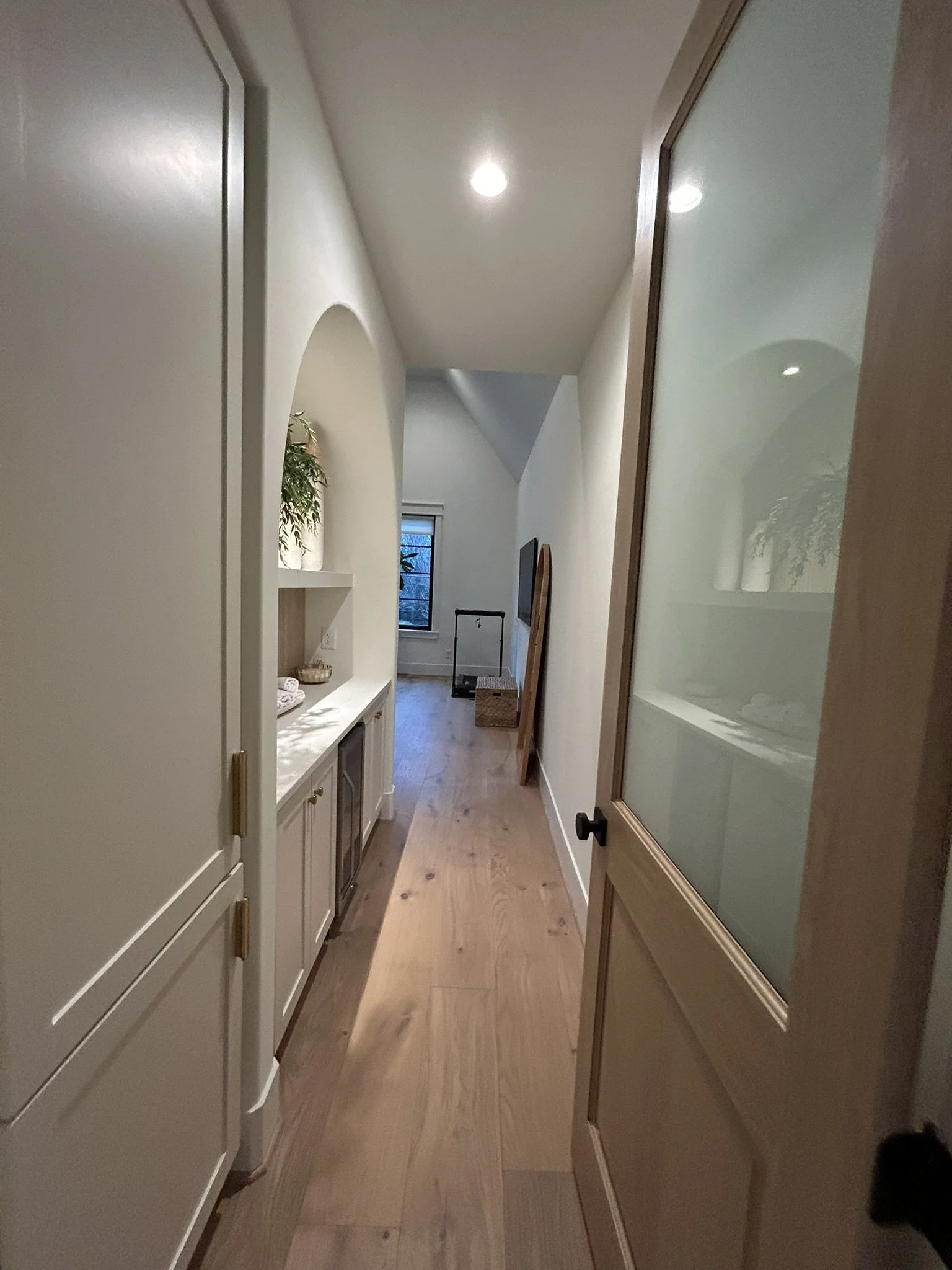 A residential hallway with wooden flooring, white walls, and built-in cabinets on the left. A window at the end of the hall reveals an outdoor scene. On the right, a wooden door with frosted glass panels is partially open.