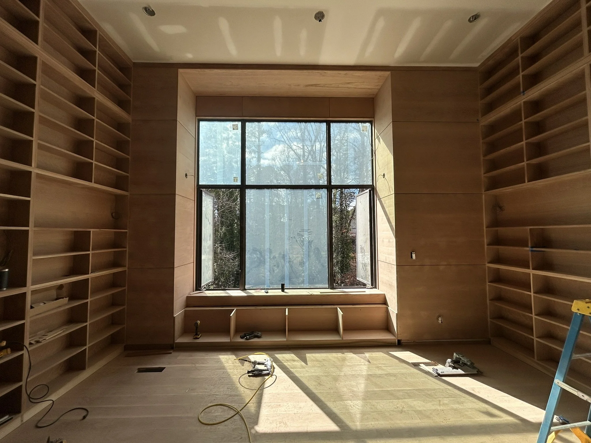 A room under construction with built-in wooden bookshelves on both sides, large window at the back letting in natural light, tools and cables on the floor, and a blue ladder on the right side.
