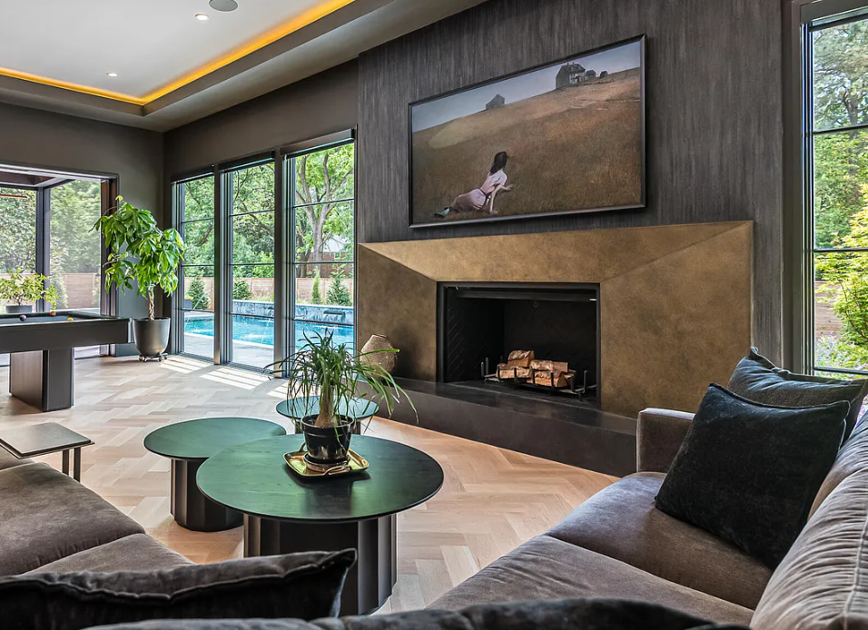 Modern living room with large windows, gray sofa, black cushions, a fireplace with a wooden mantel, a painting above the fireplace, a large potted plant, and round black coffee tables.