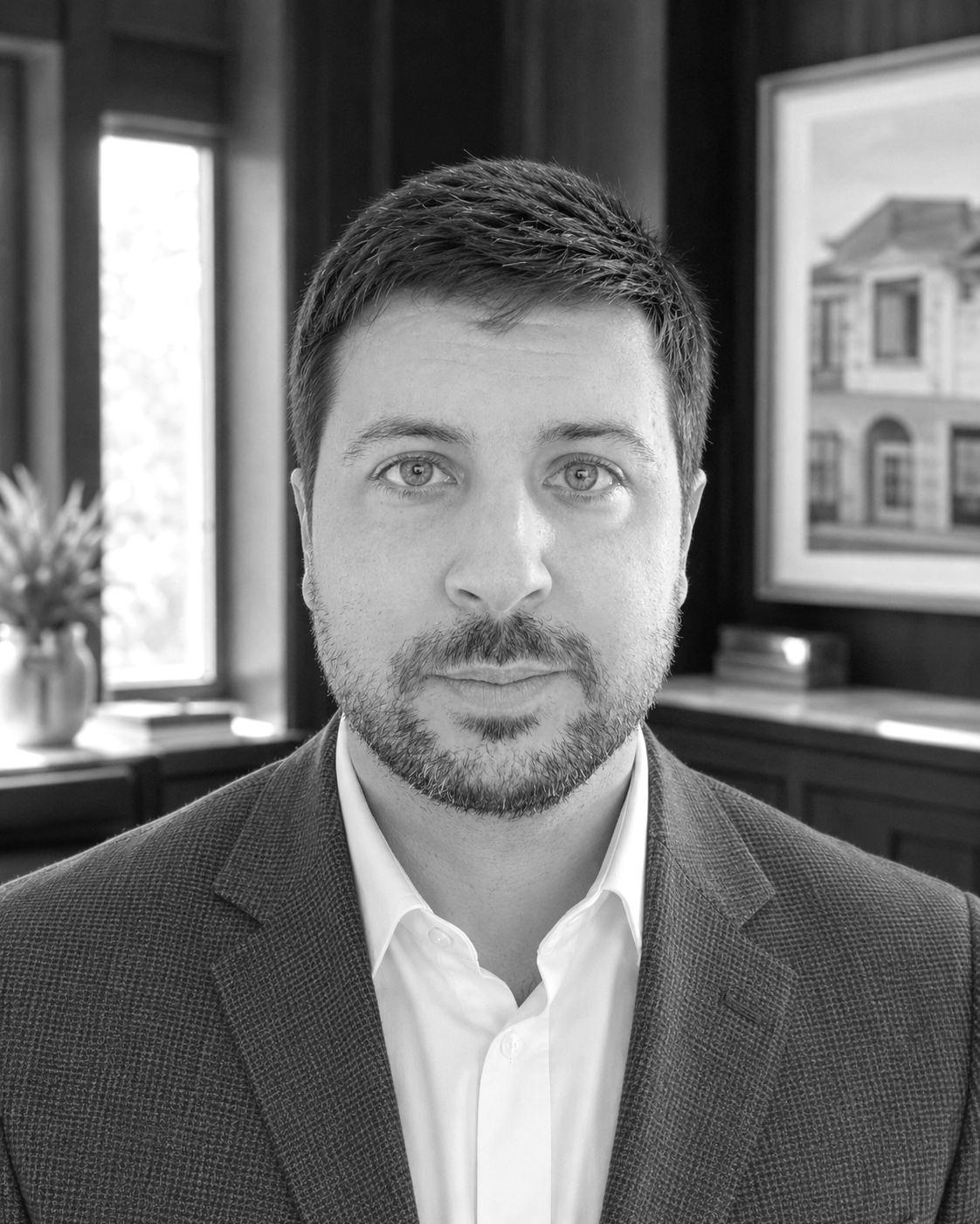 A black-and-white portrait of a man with short dark hair and a beard, wearing a suit jacket and a collared shirt, indoors with windows and framed pictures in the background.