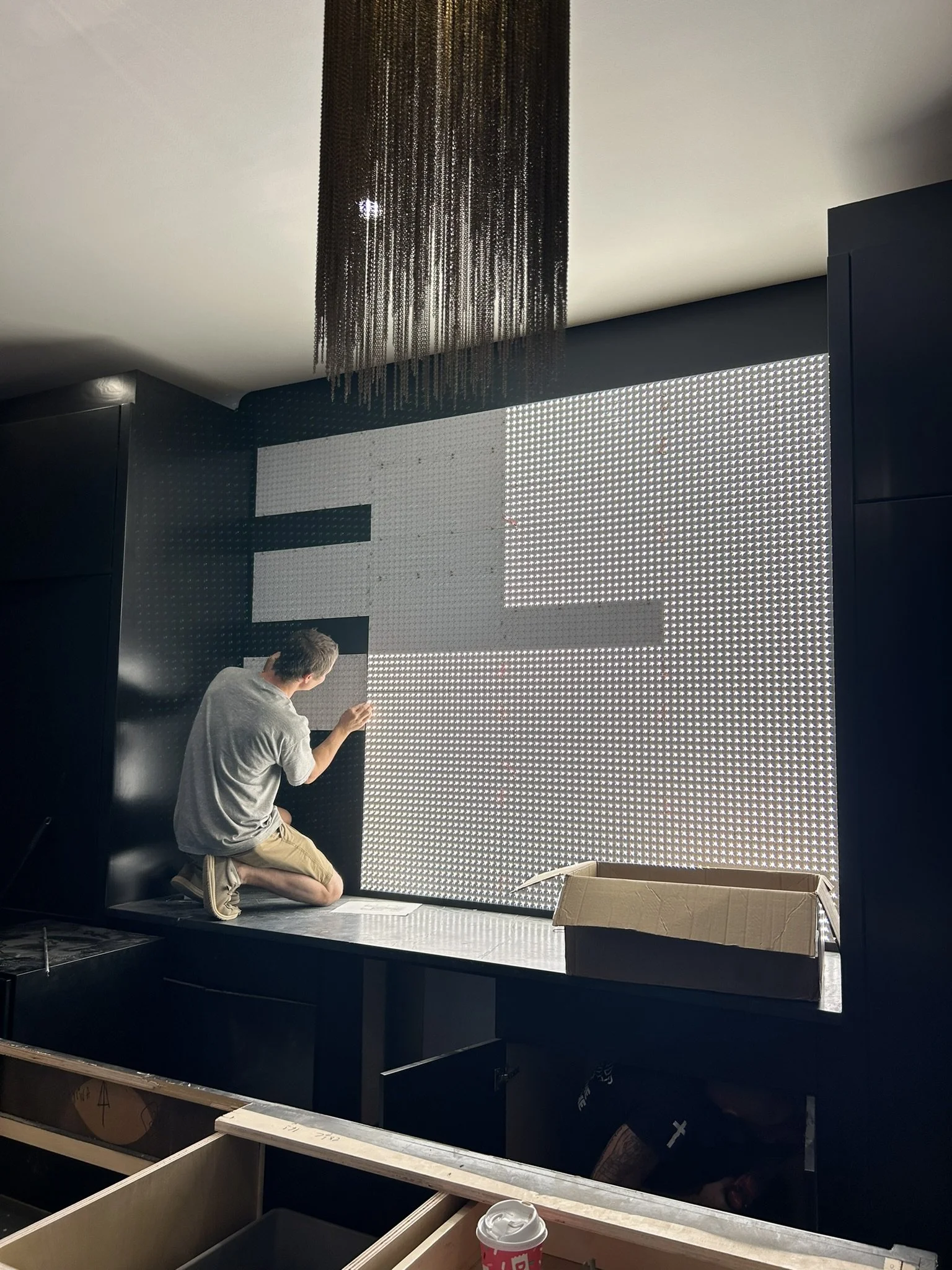 A person kneeling on a bench working on a large illuminated grid wall with a black background. The person is wearing a gray shirt and khaki shorts, and there is an open cardboard box and a coffee cup in the foreground.