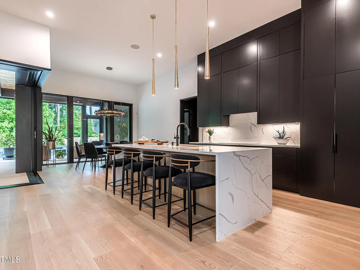 Modern kitchen with white marble island, black cabinetry, and light wood flooring, featuring pendant lights and large windows with a view of greenery.