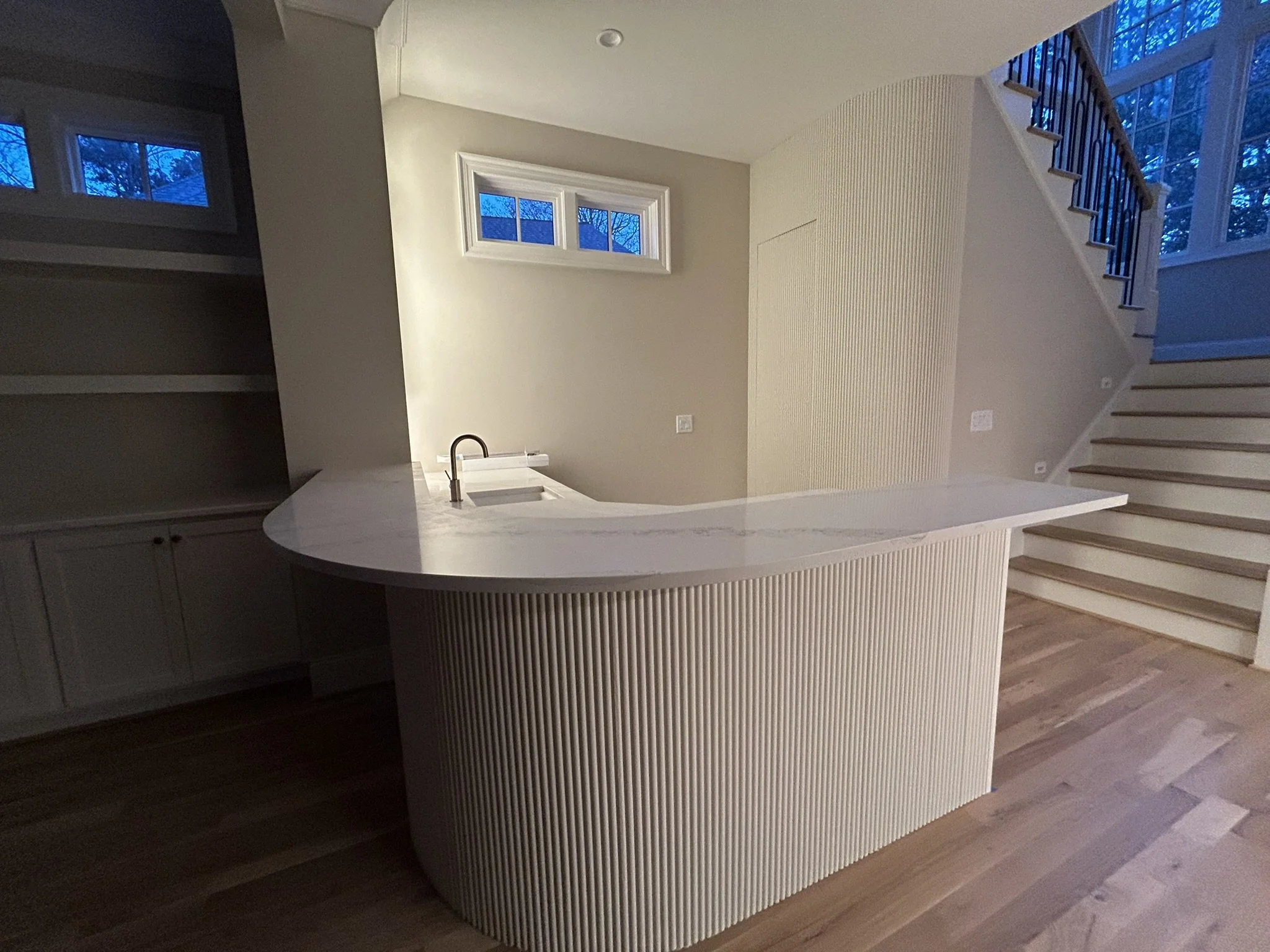 Interior view of a modern home with a staircase, large windows showing trees outside, and a curved kitchen island with a sink.