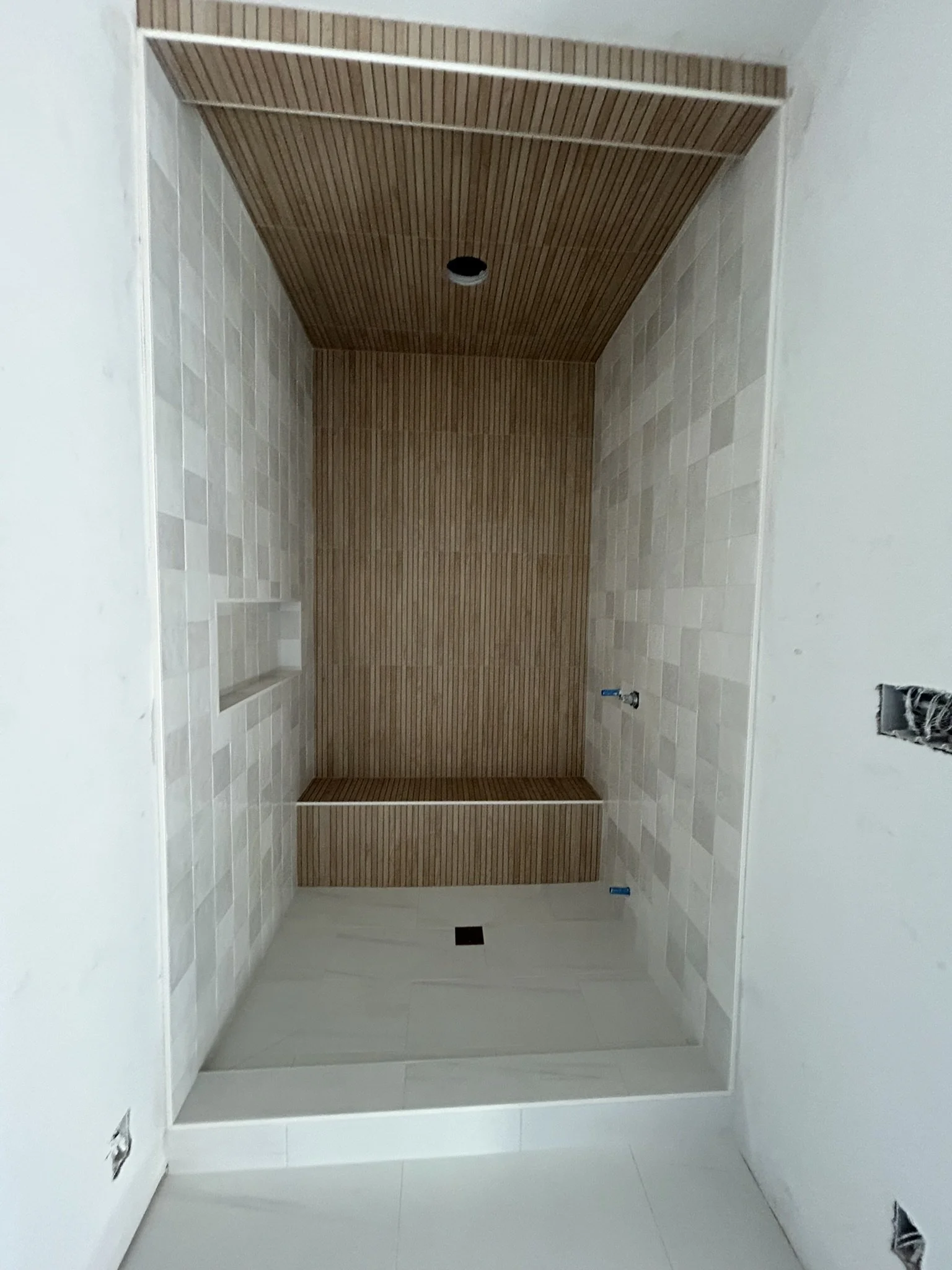 An interior space under construction with white-tiled walls, a wooden ceiling with narrow slats, and an opening in the wall framed with wood. There are electrical boxes and outlets visible on the walls.