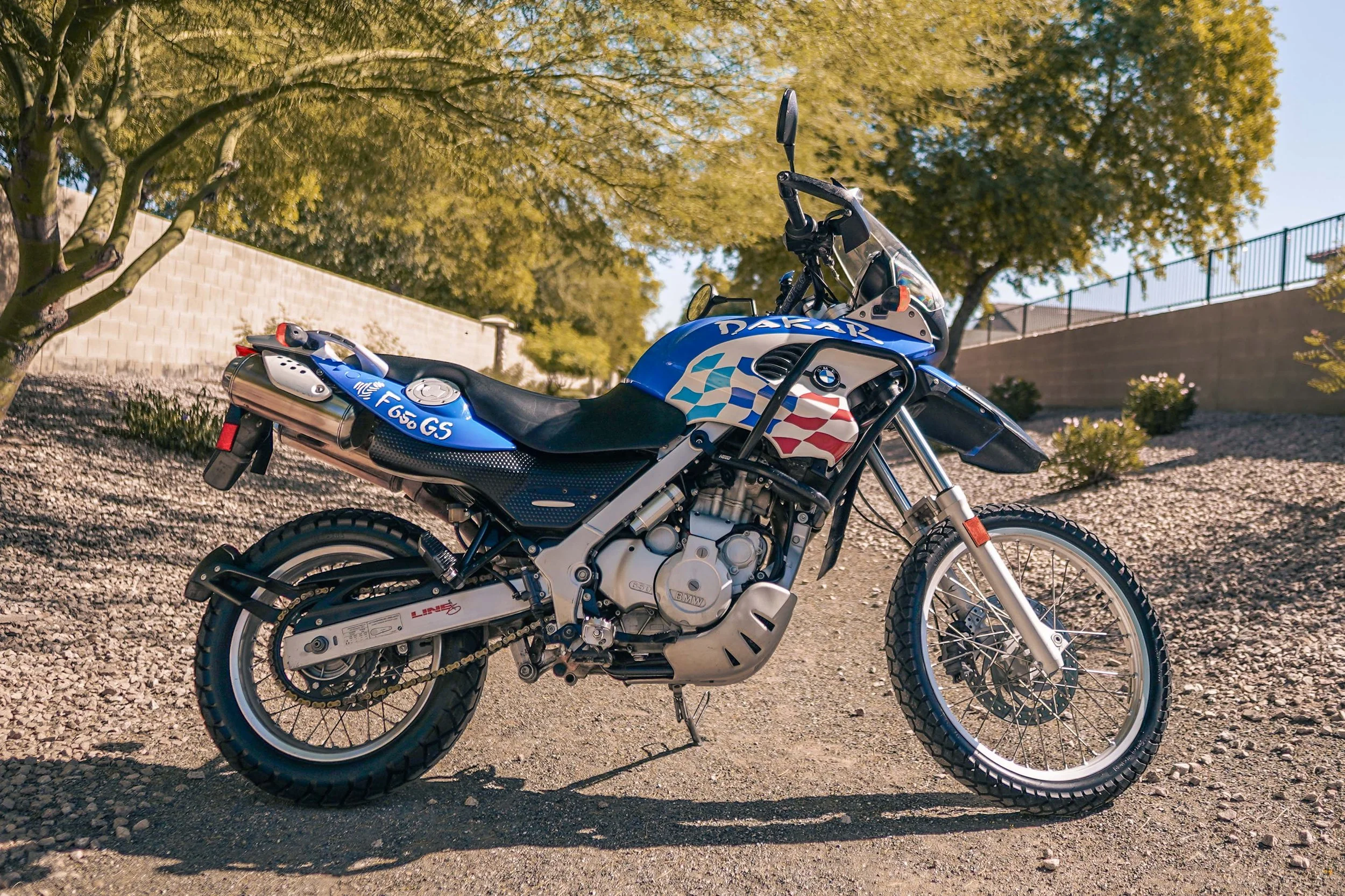 Blue BMW motorcycle with Dakar and racing decals parked on gravel, trees with yellow-green leaves in the background.