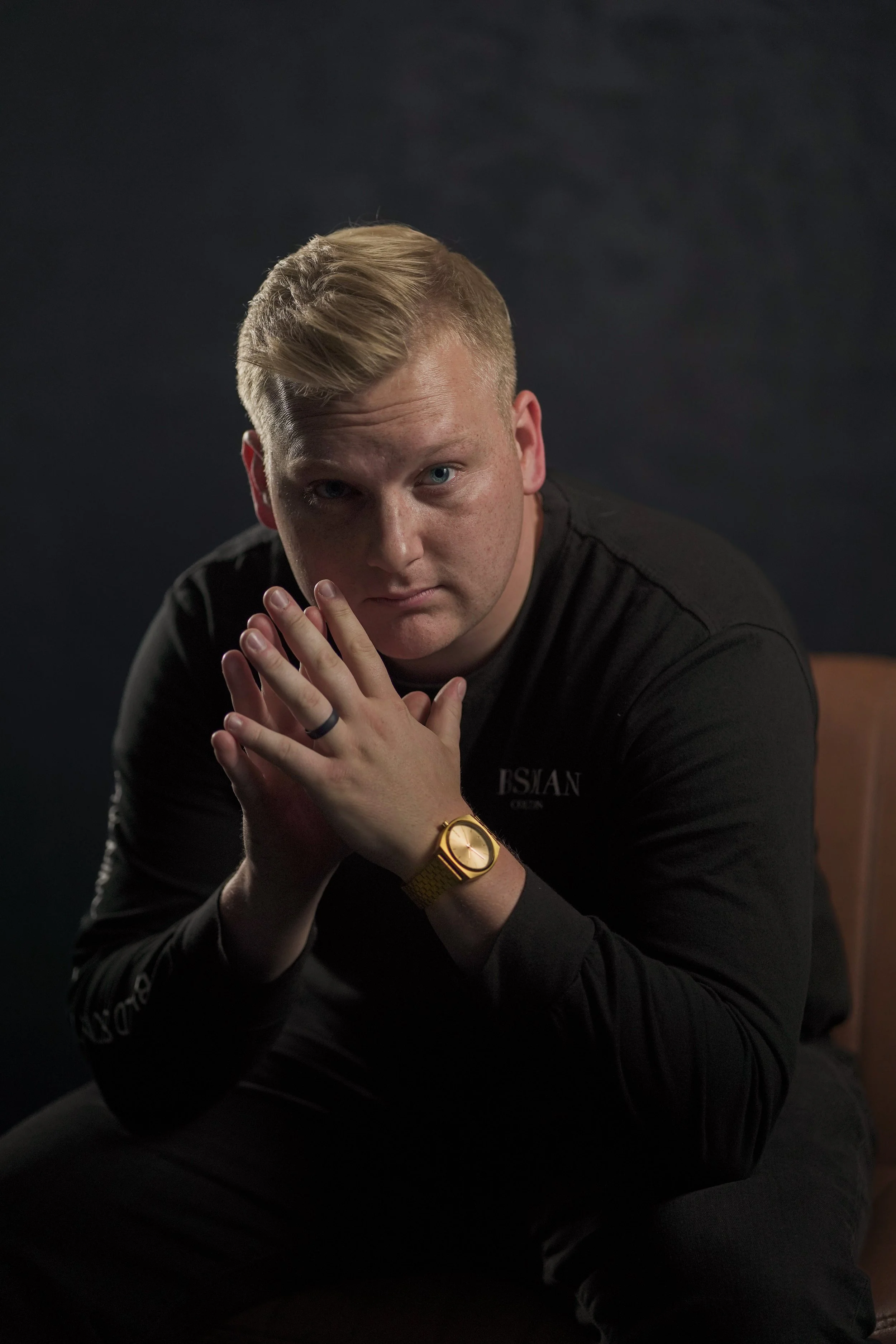 A young man with blonde hair and blue eyes, wearing a black long-sleeve shirt with white text, a gold wristwatch, and a black ring, sitting against a dark background, looking directly into the camera with his hands clasped together.
