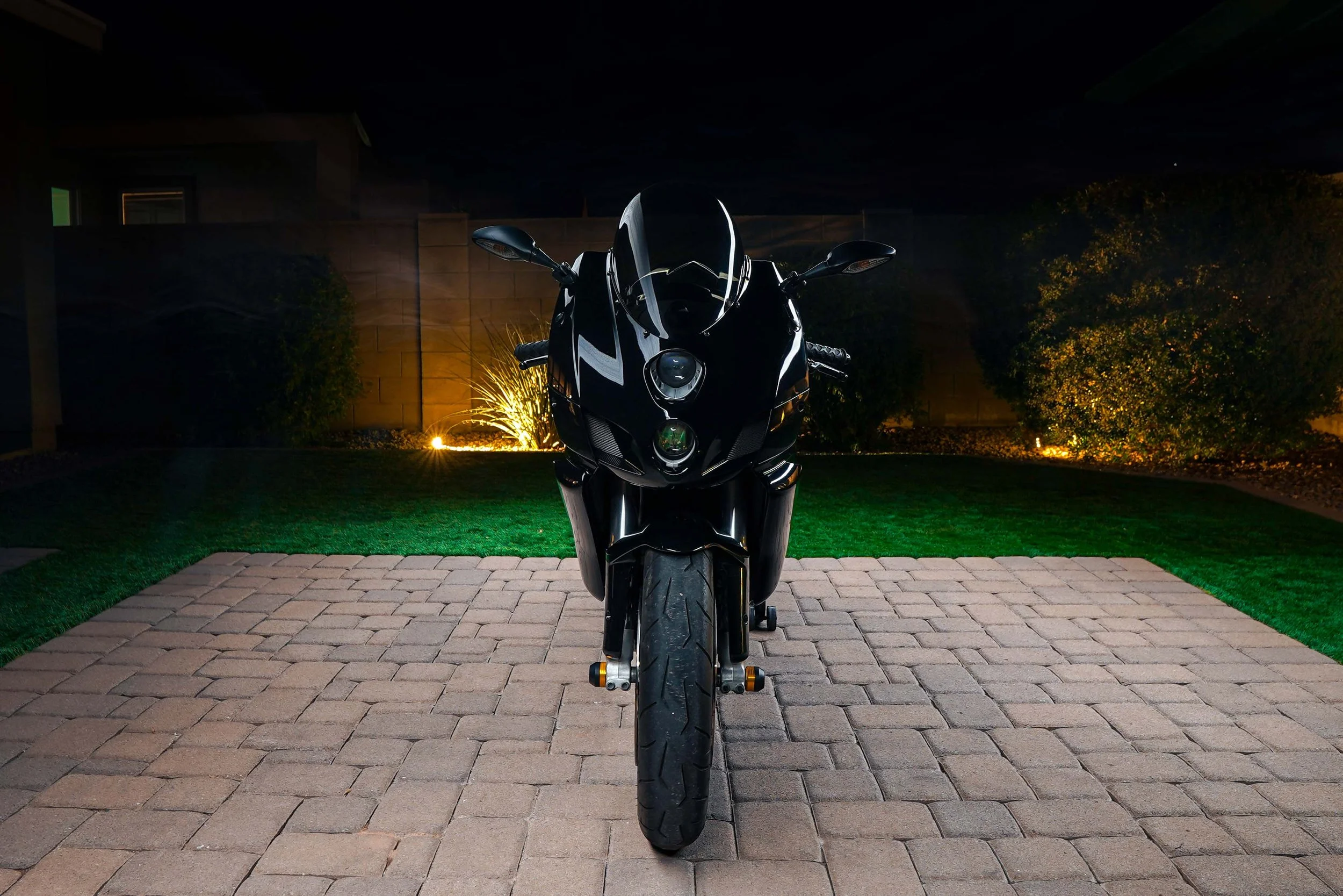 Front view of a black motorcycle parked on a paved surface at night, illuminated by ground lights with a backyard and dark sky in the background.