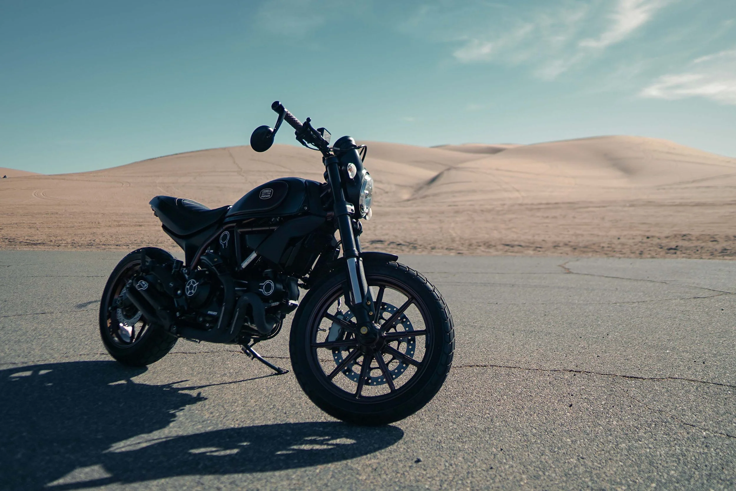 A black motorcycle parked on a paved road in a desert landscape with sandy dunes and a blue sky.