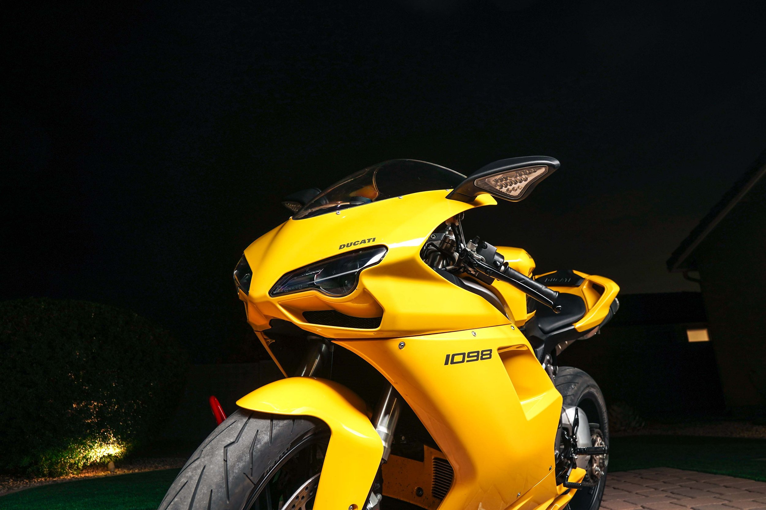 Yellow Ducati motorcycle parked outdoors at night, illuminated with black sky and some greenery in the background.