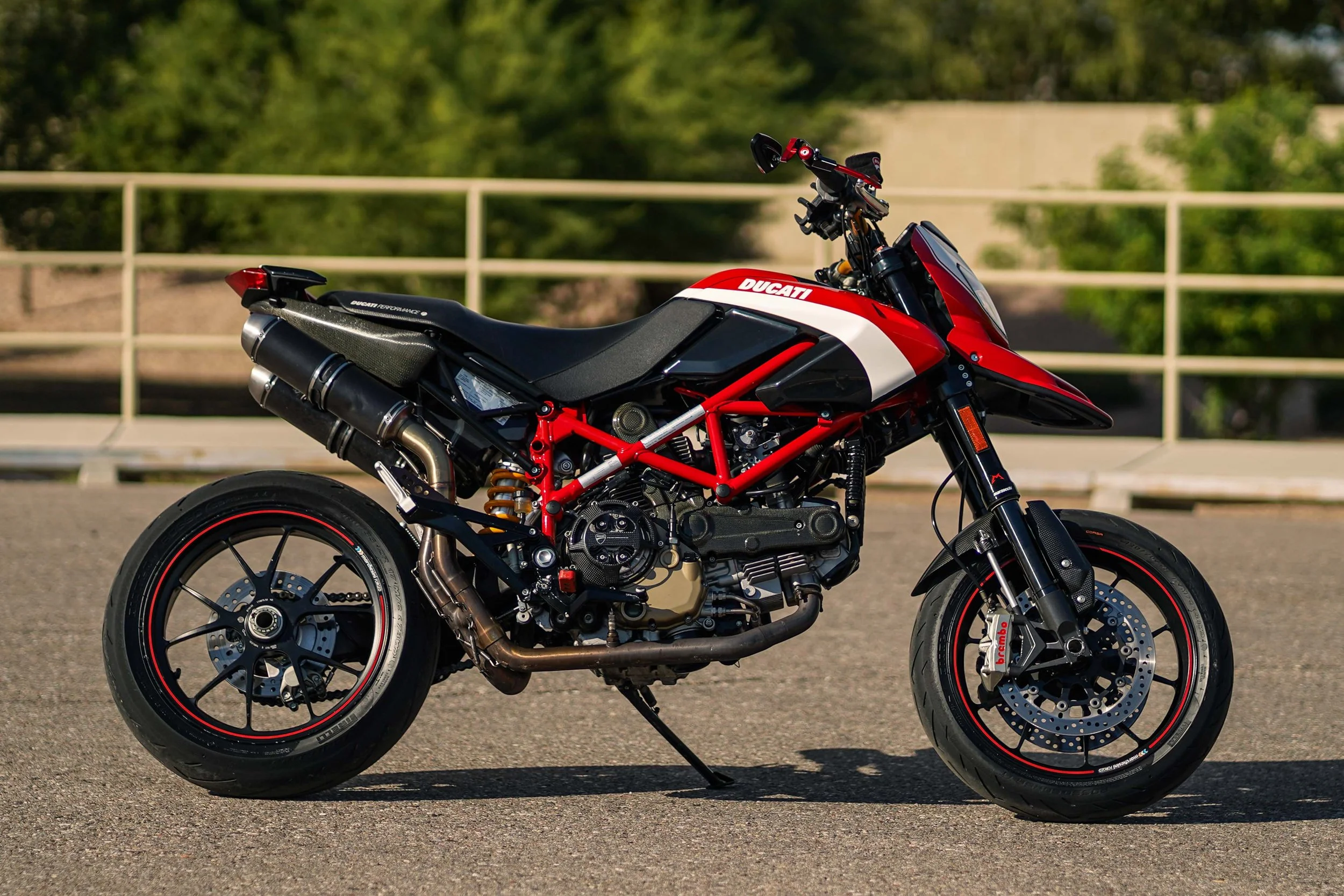 A red and black Ducati motorcycle parked on a paved surface with a background of trees and a white fence.