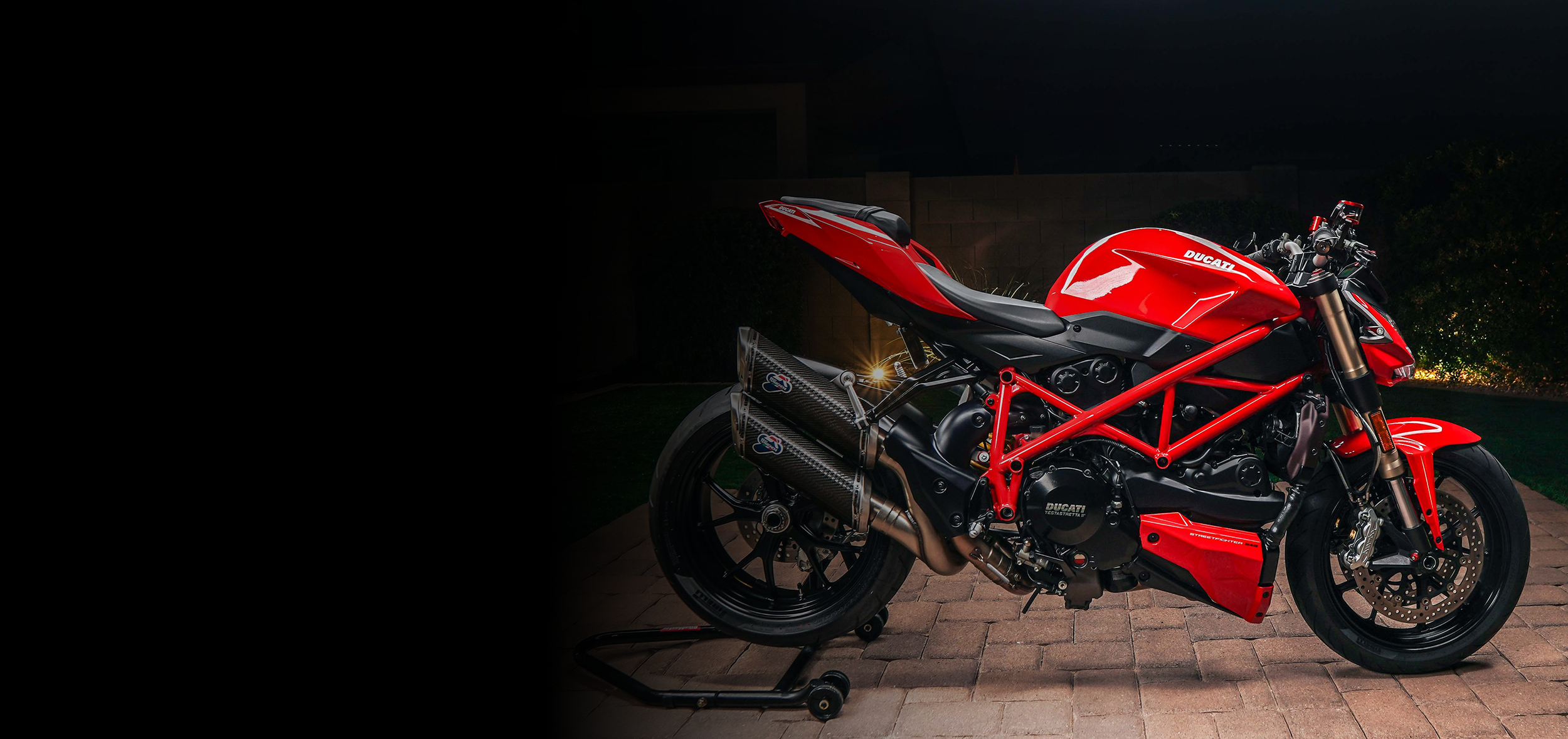Red Ducati sport motorcycle parked on brick pavement at night, with dark background and some greenery.