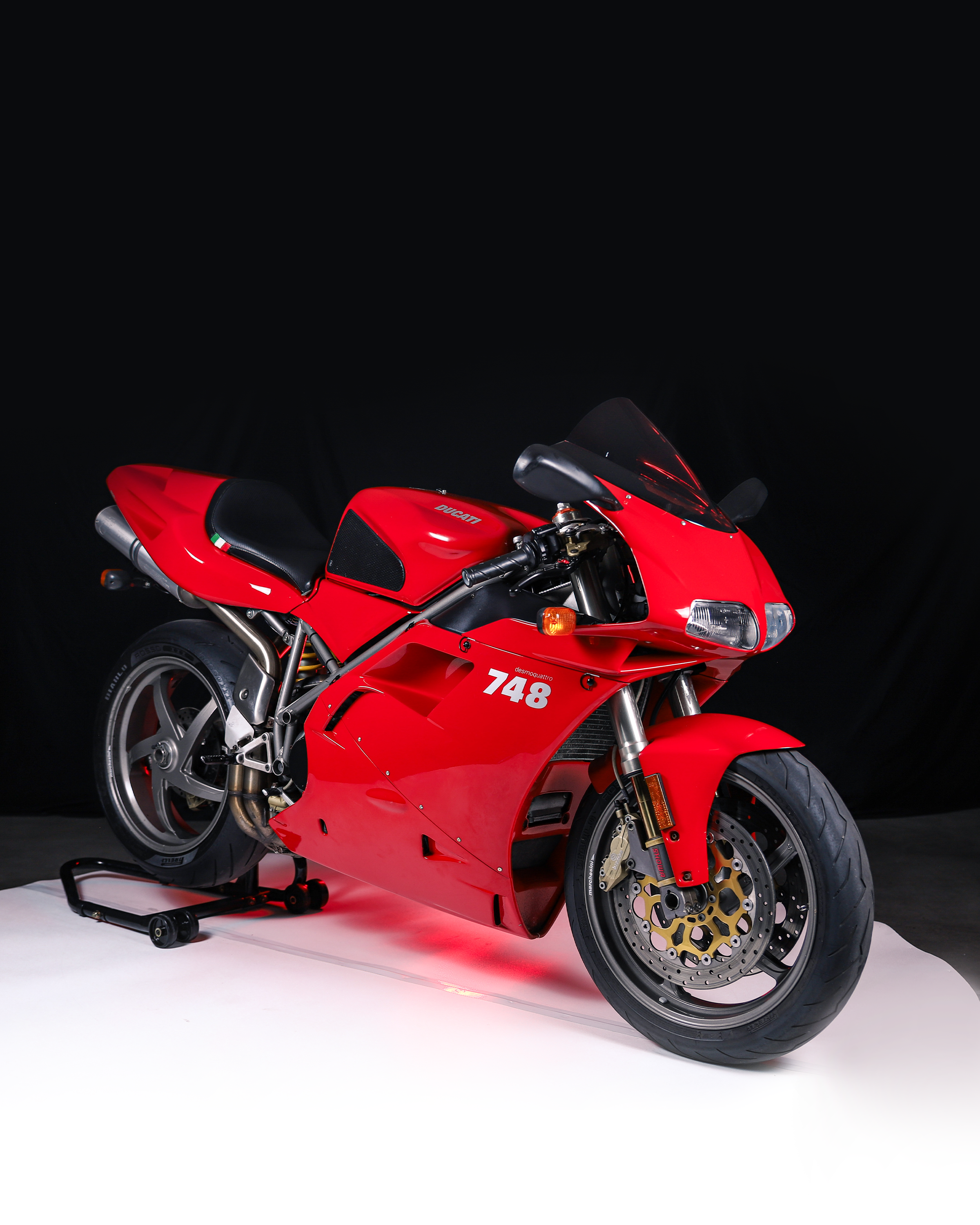 A red Ducati 748 sport motorcycle with a black seat and front windshield, displayed on a white platform against a black background.