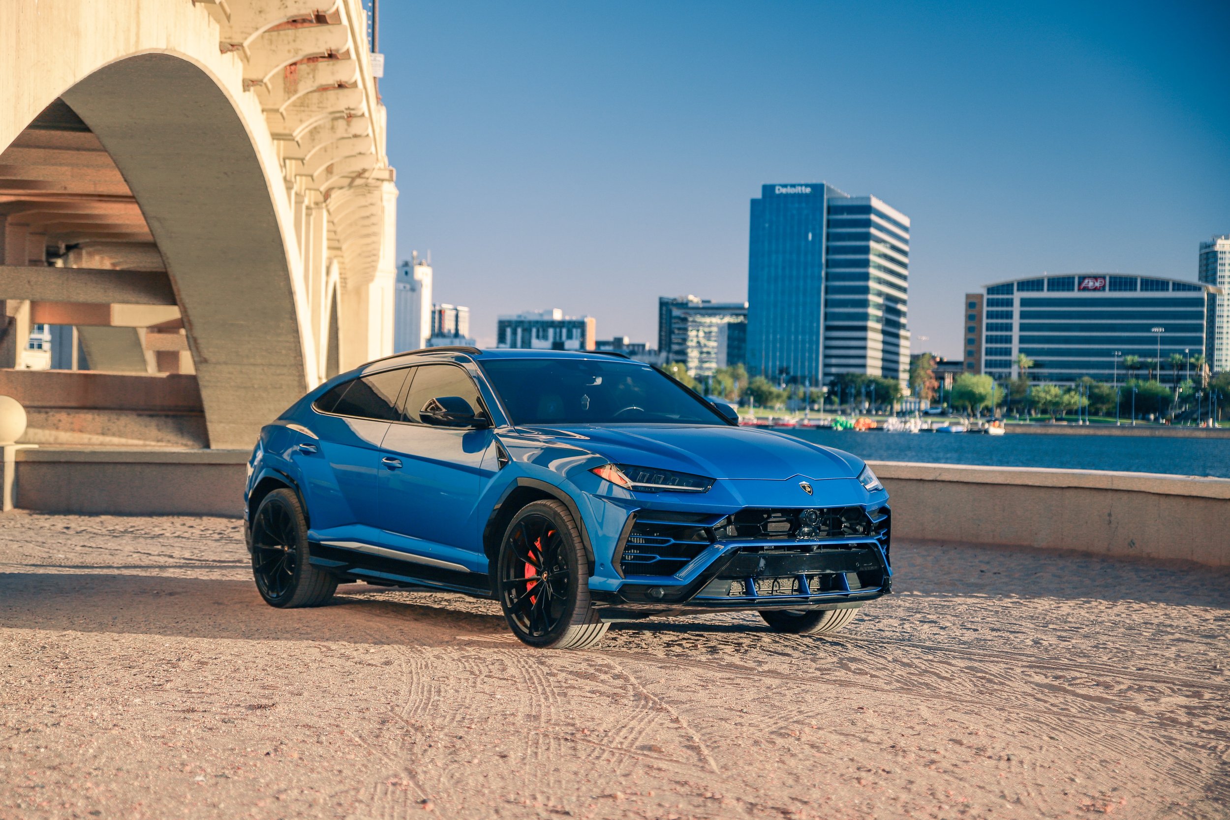 A blue Lamborghini parked on a sandy area near a waterfront with city buildings in the background and a clear blue sky.
