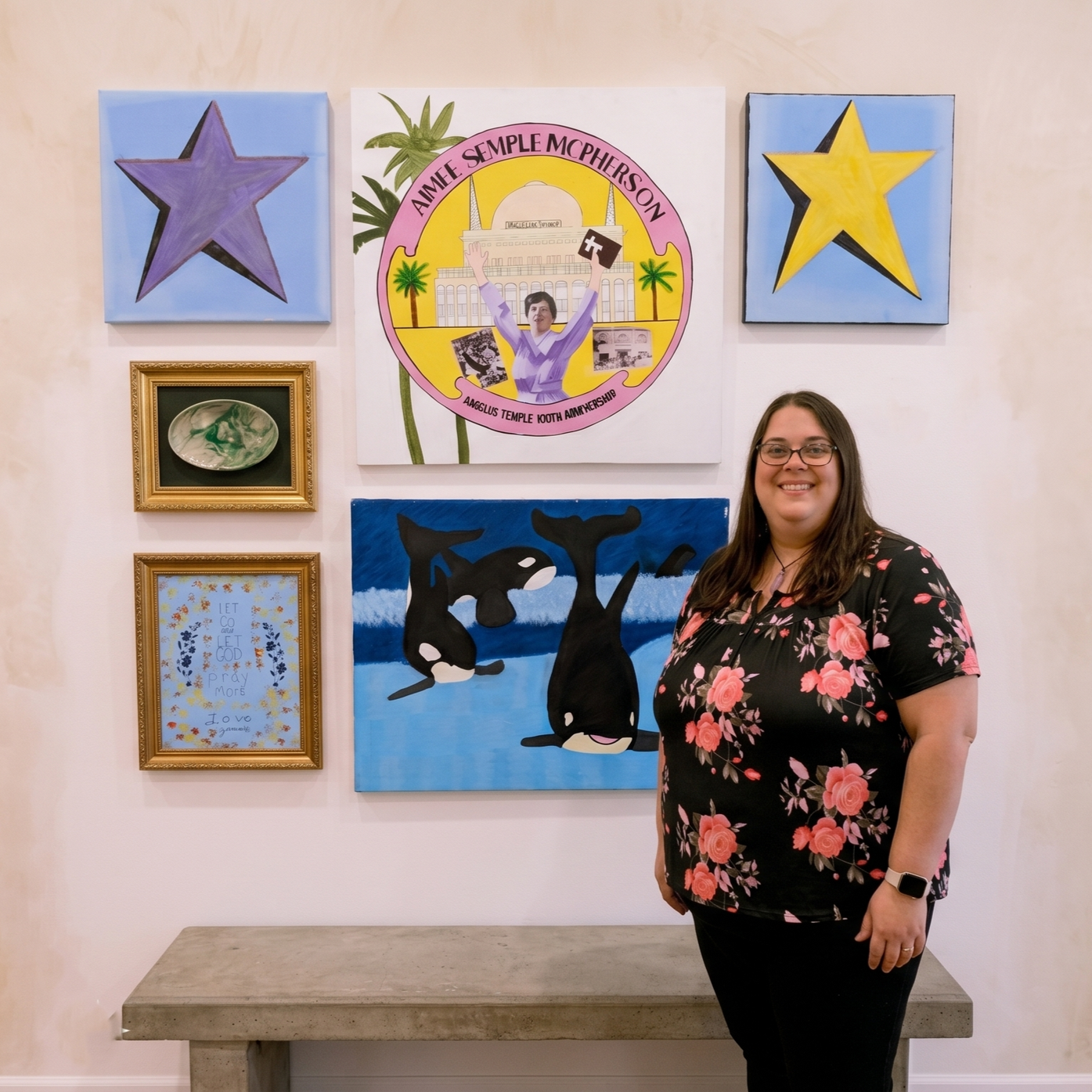 A woman smiling and standing in front of a wall with various framed artwork and paintings, including a large piece with a woman holding a flag and a building in the background, and two star paintings, one purple and one yellow. There are also paintings of orcas and a framed quote that says 'Let go and let God pray more'.