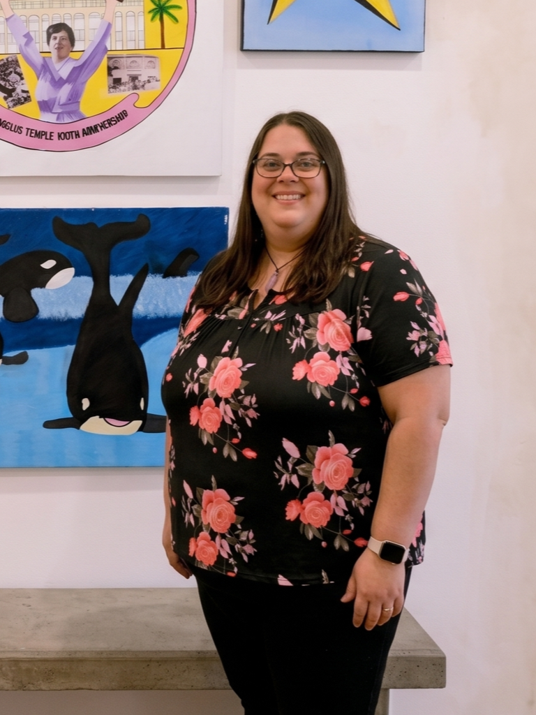 A woman with glasses and brown hair standing indoors in front of colorful artwork, wearing a black floral shirt with pink roses, black pants, and a smartwatch.
