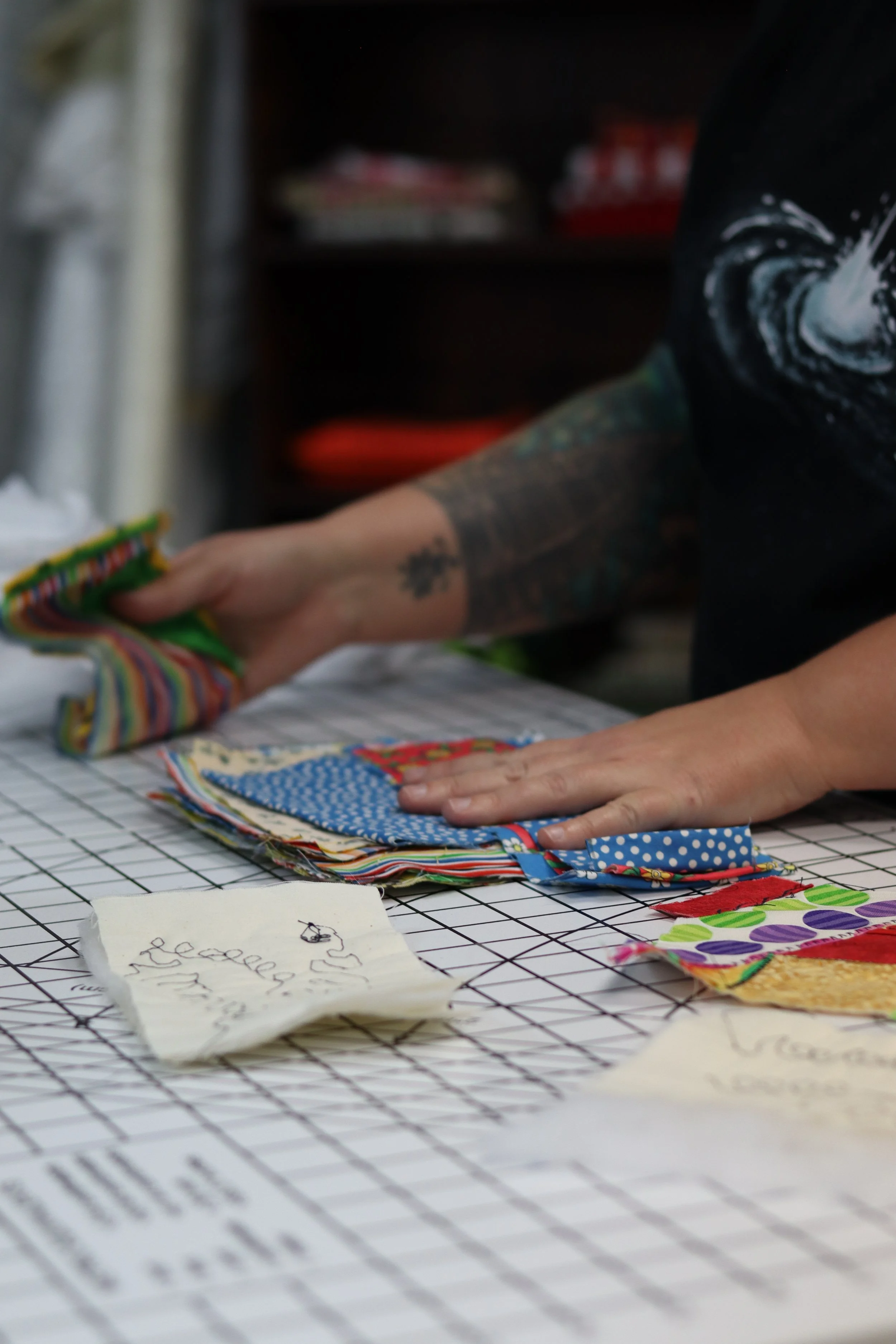 Close-up of a person's hand arranging colorful fabric scraps on a grid-patterned surface, with handwritten notes on paper in the foreground.