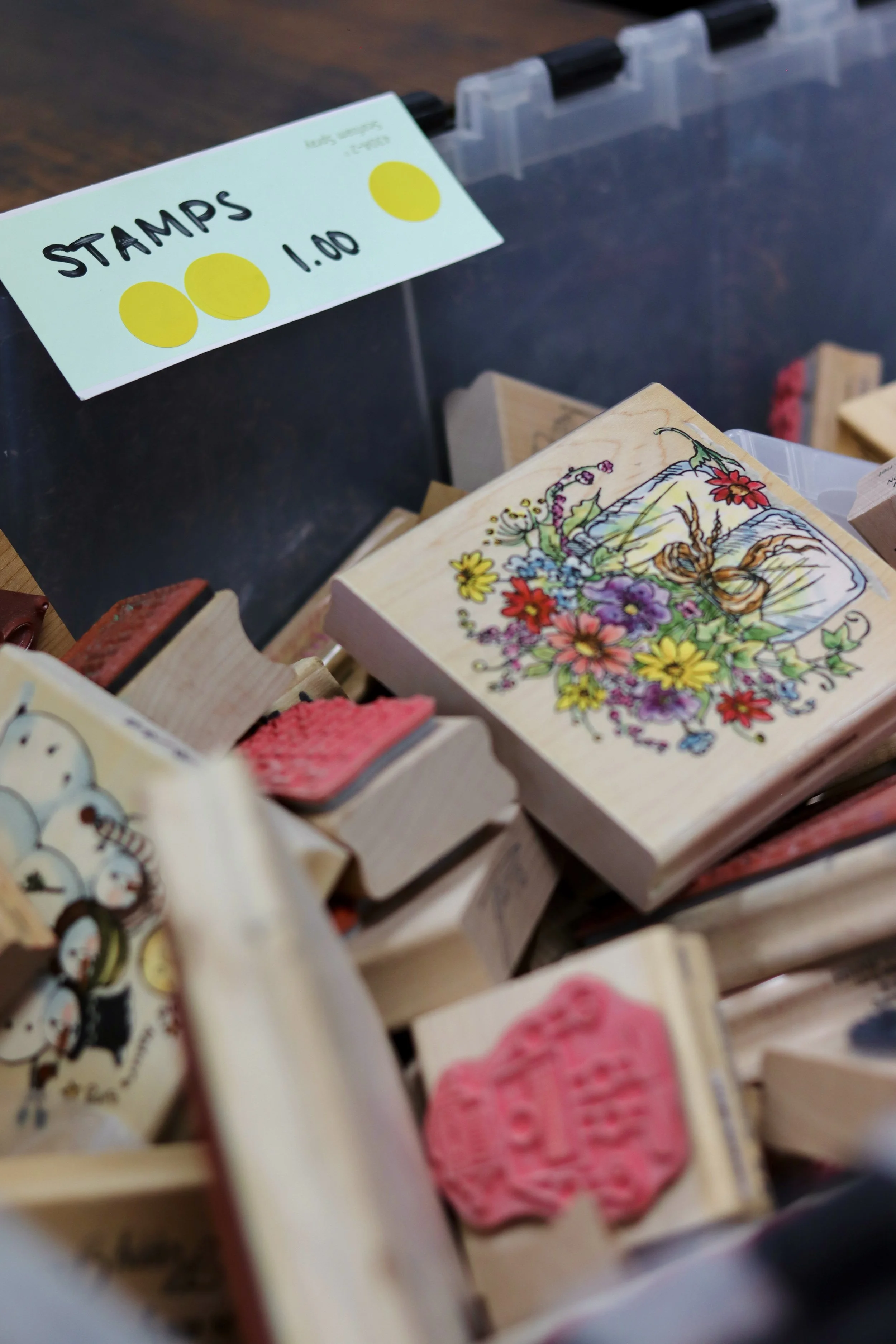 A display of wooden stamps with various colorful designs and stamps on a store shelf, labeled with a sign that says 'Stamps 1.00' featuring three yellow circles.