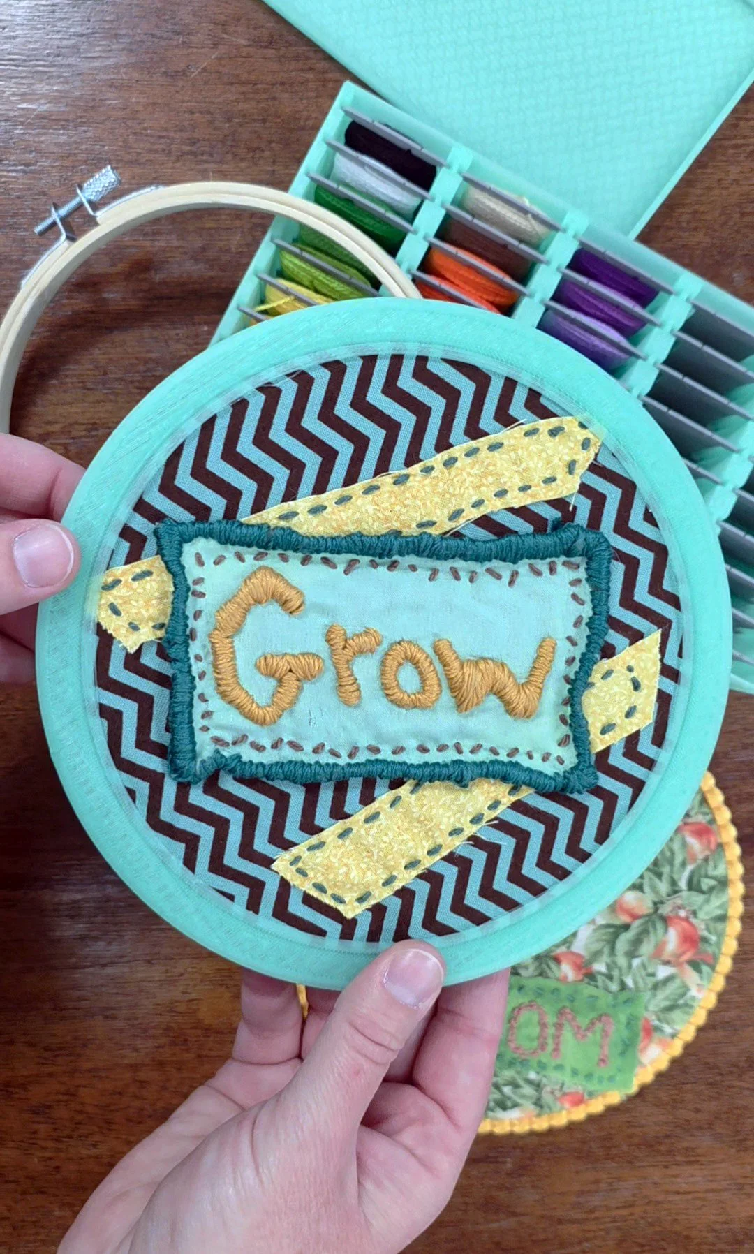 A person's hands holding a circular fabric piece with the word 'Grow' embroidered in yellow thread. The fabric is decorated with yellow and teal patterns and is part of a collection of embroidery hoop projects. In the background, there is a box of colorful embroidery floss and another embroidered piece on a wooden surface.