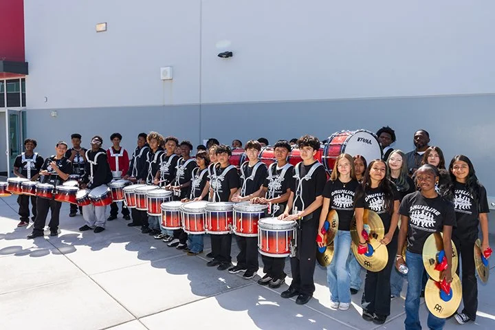 Group of students in black shirts holding musical instruments, including drums and cymbals, standing outside against a plain wall.