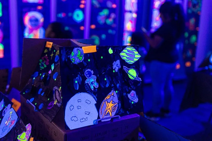 Box decorated with glow-in-the-dark space-themed drawings, including planets, rockets, and astronauts, with children in the background at an indoor glow-in-the-dark party.
