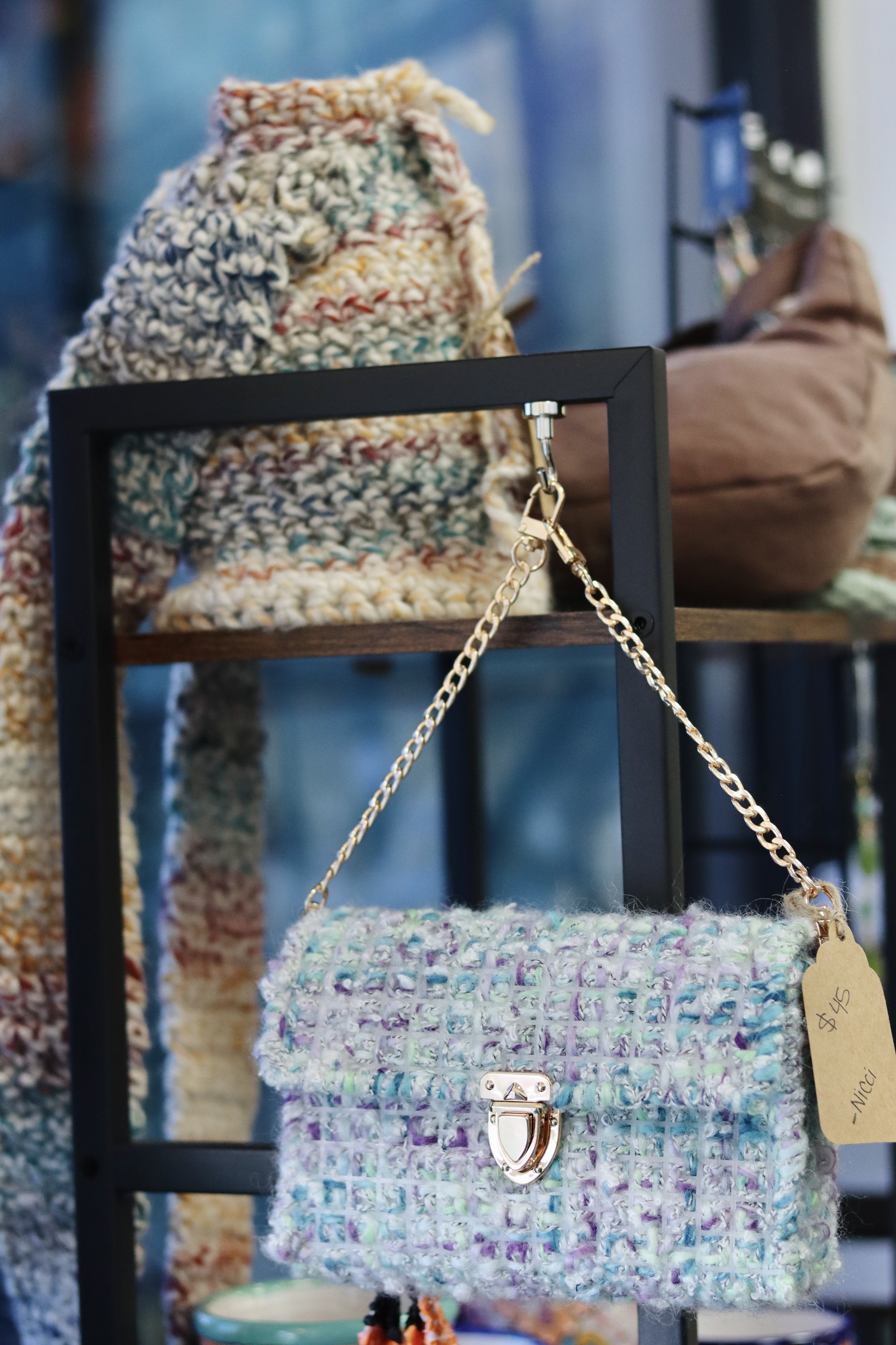 Close-up of a multicolored textured handbag with a chain strap displayed on a black metal stand at a craft or artisan market.