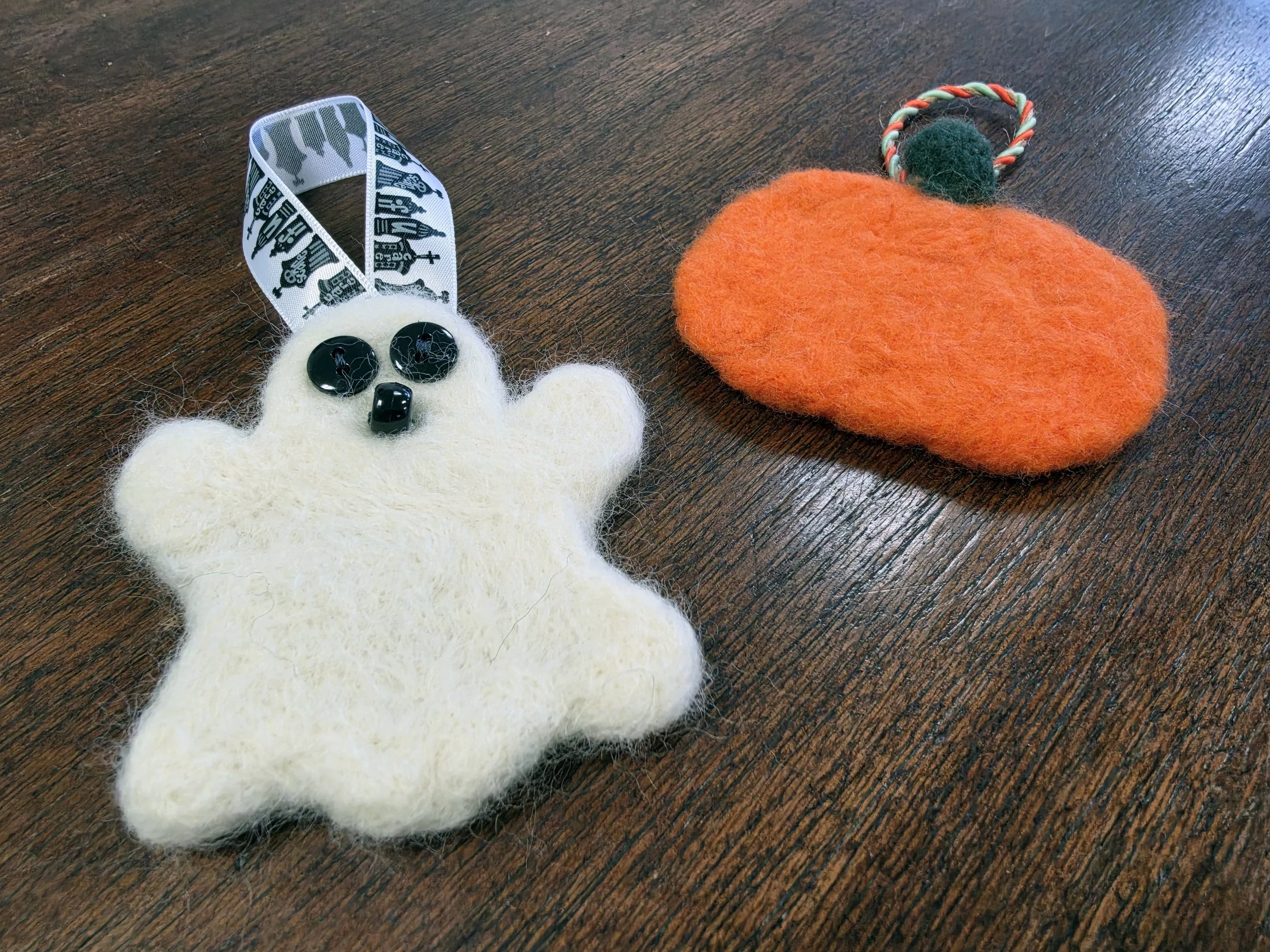 Felted wool craft items, including a ghost with black eyes and buttons, and a bright orange pumpkin, on a wooden surface.