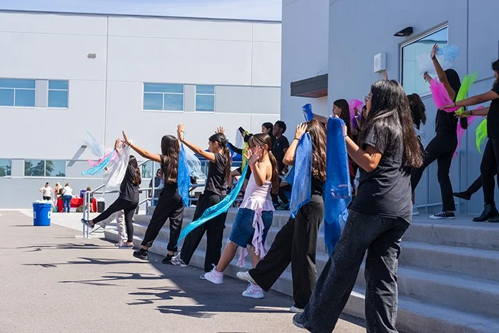 A group of young women performing a dance or cheer routine outside a modern building, holding colorful scarves or flags.