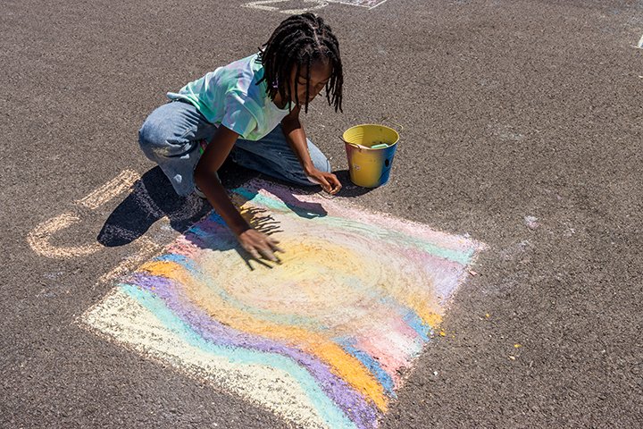 A young girl is kneeling on the asphalt, creating a colorful chalk drawing on the ground. She has a bucket of chalk nearby.
