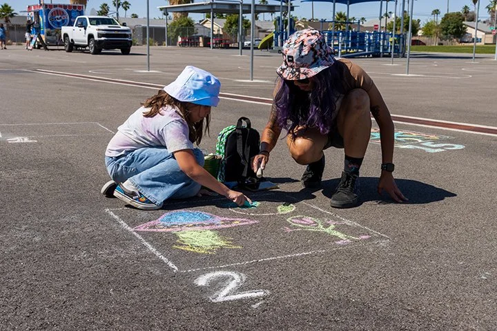 Two girls drawing hopscotch and colorful chalk art on a parking lot pavement, with a pickup truck and playground equipment in the background on a sunny day.