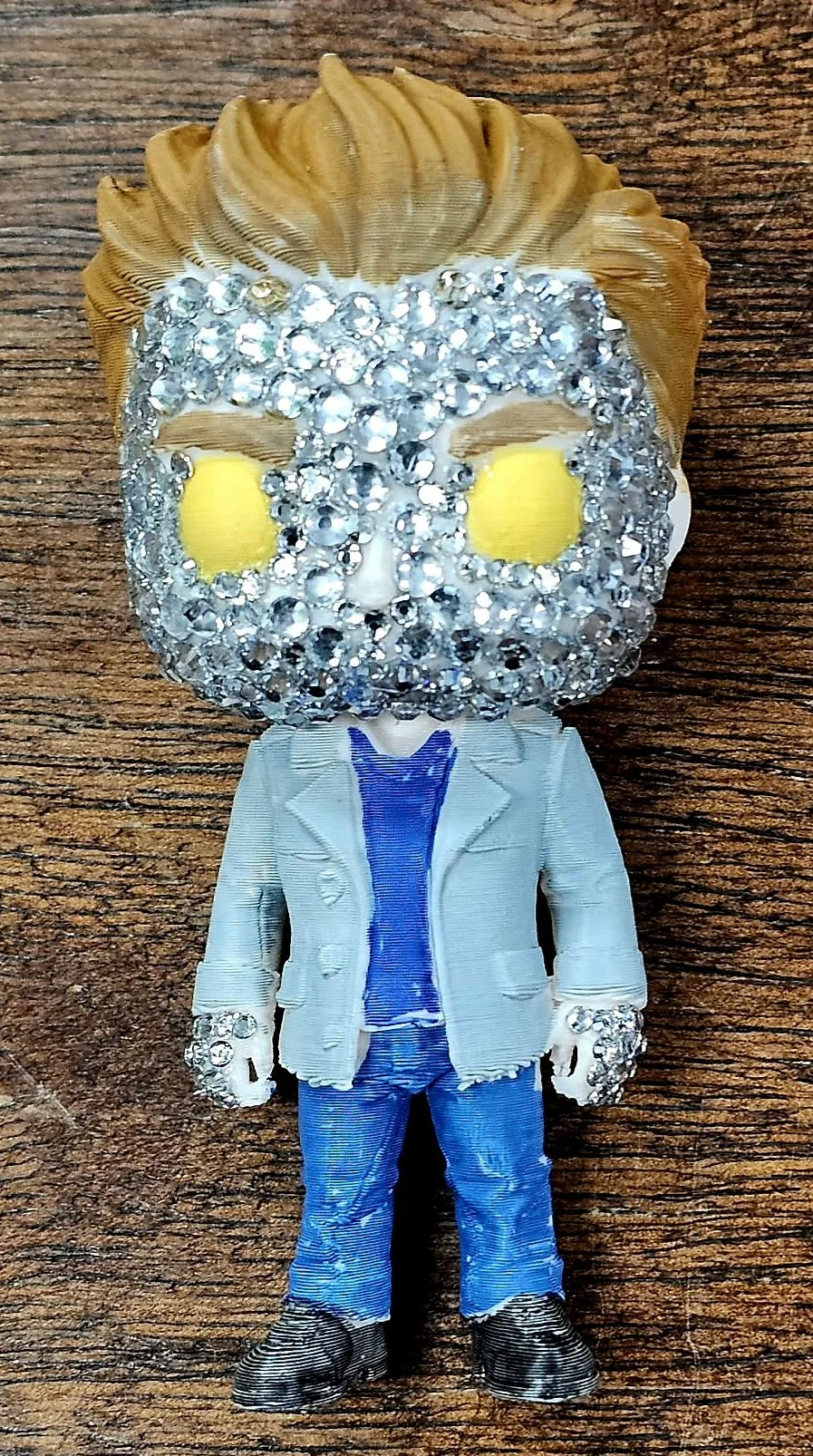 A plush toy with a large head decorated with silver rhinestones, resembling a stylized person with blond hair. The toy is dressed in a white blazer, blue jeans, and black shoes, standing on a wooden surface.
